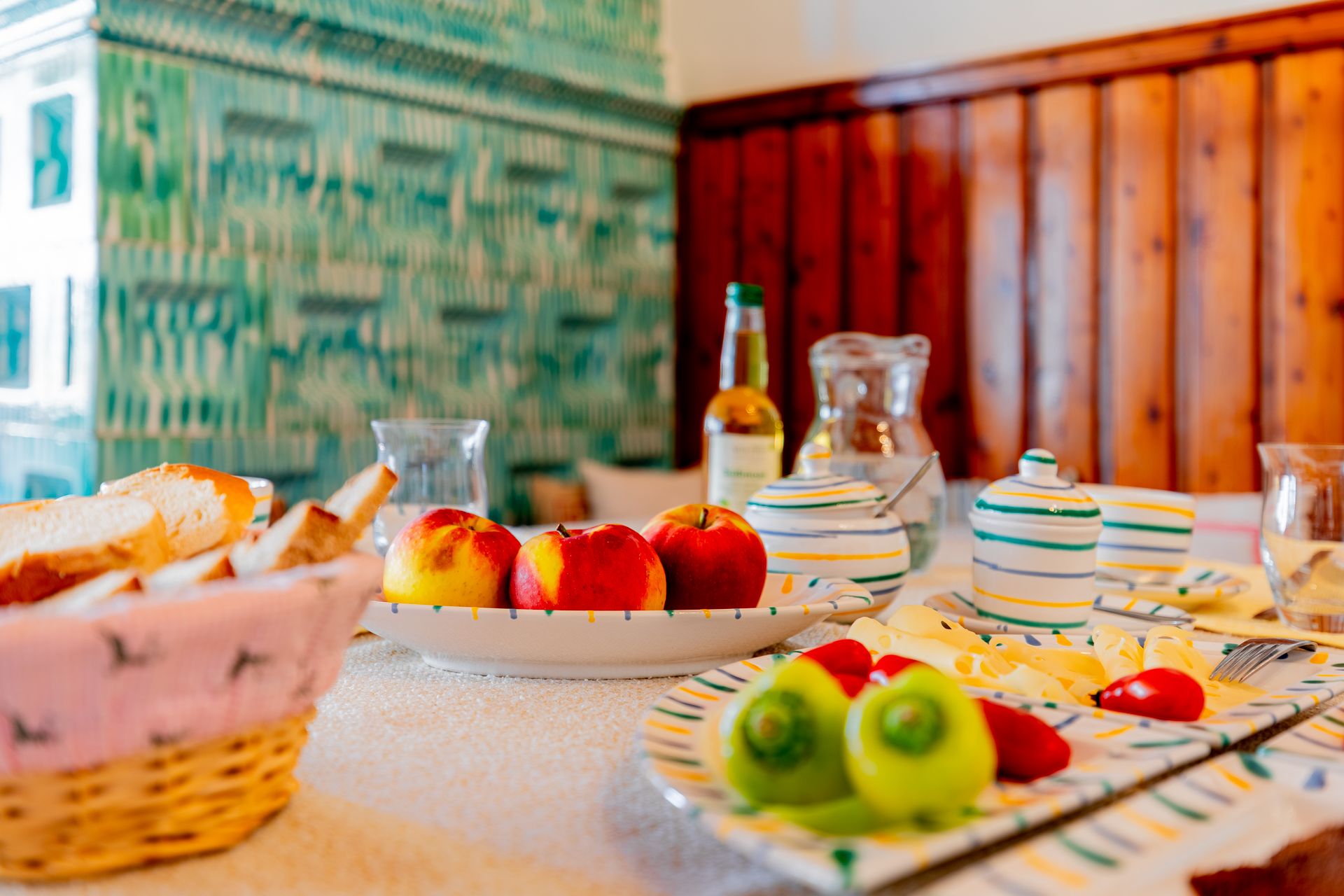A table laid with fruit, bread and crockery in front of a green tiled wall.
