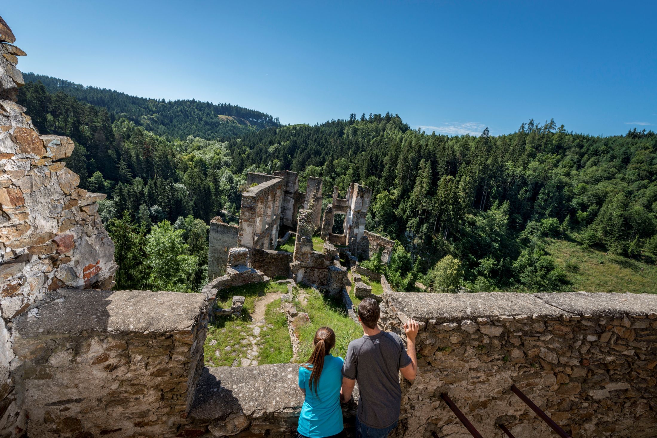 Two people look at the Kollmitz ruins from a wall, surrounded by a dense forest and blue sky.