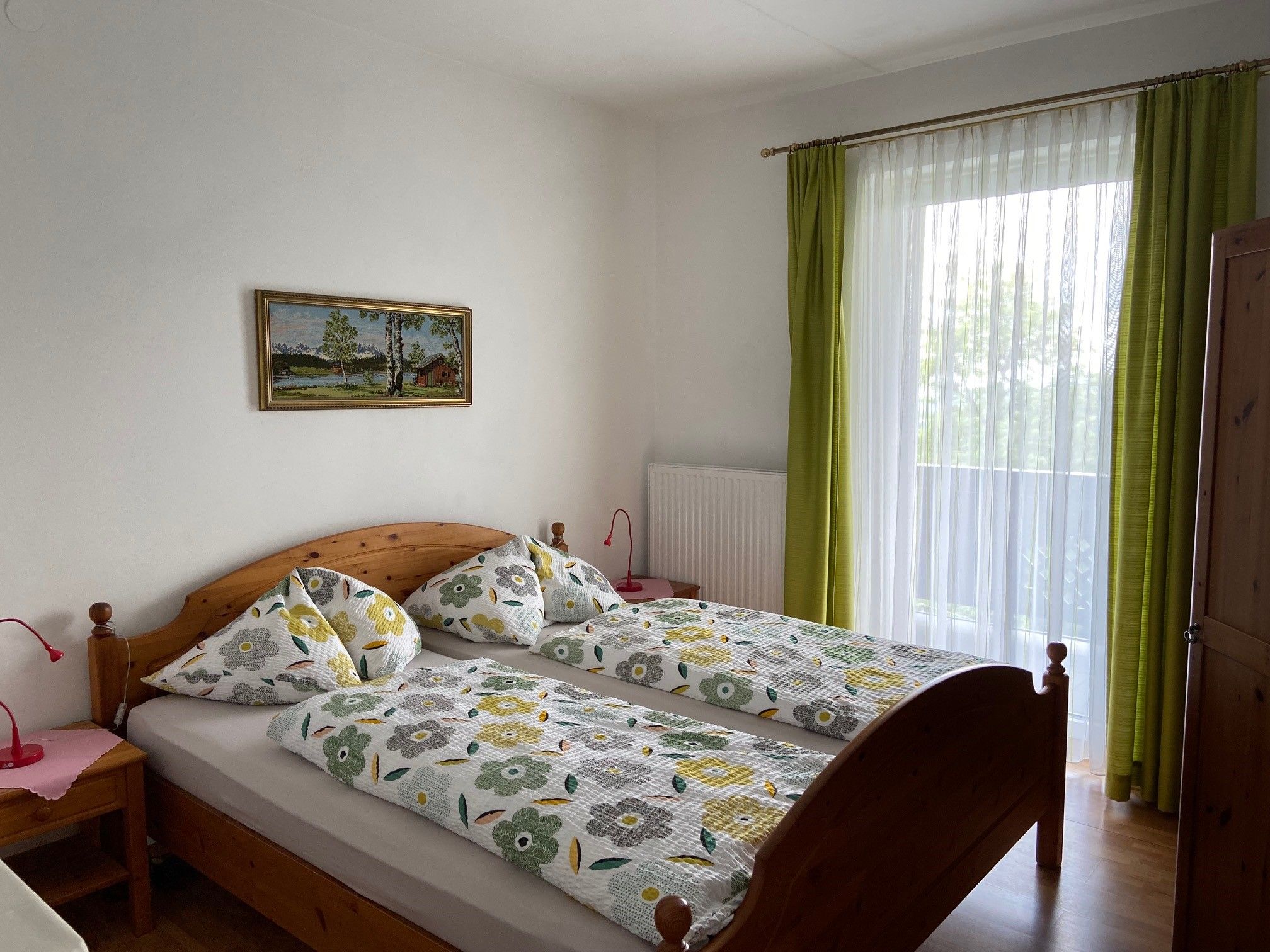 A cozy double room with wooden furniture, a large window with green curtains and a landscape painting on the wall.
