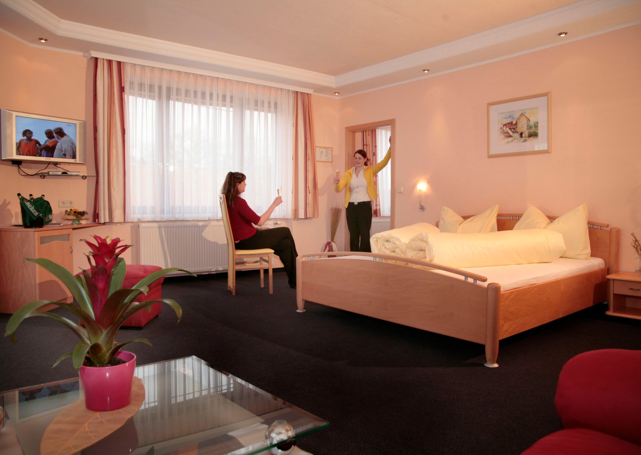 Two women in a hotel room with a double bed, one sitting, the other standing with her glass raised.