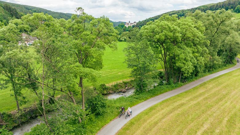 Two cyclists on a path along a river, surrounded by green trees and meadows.