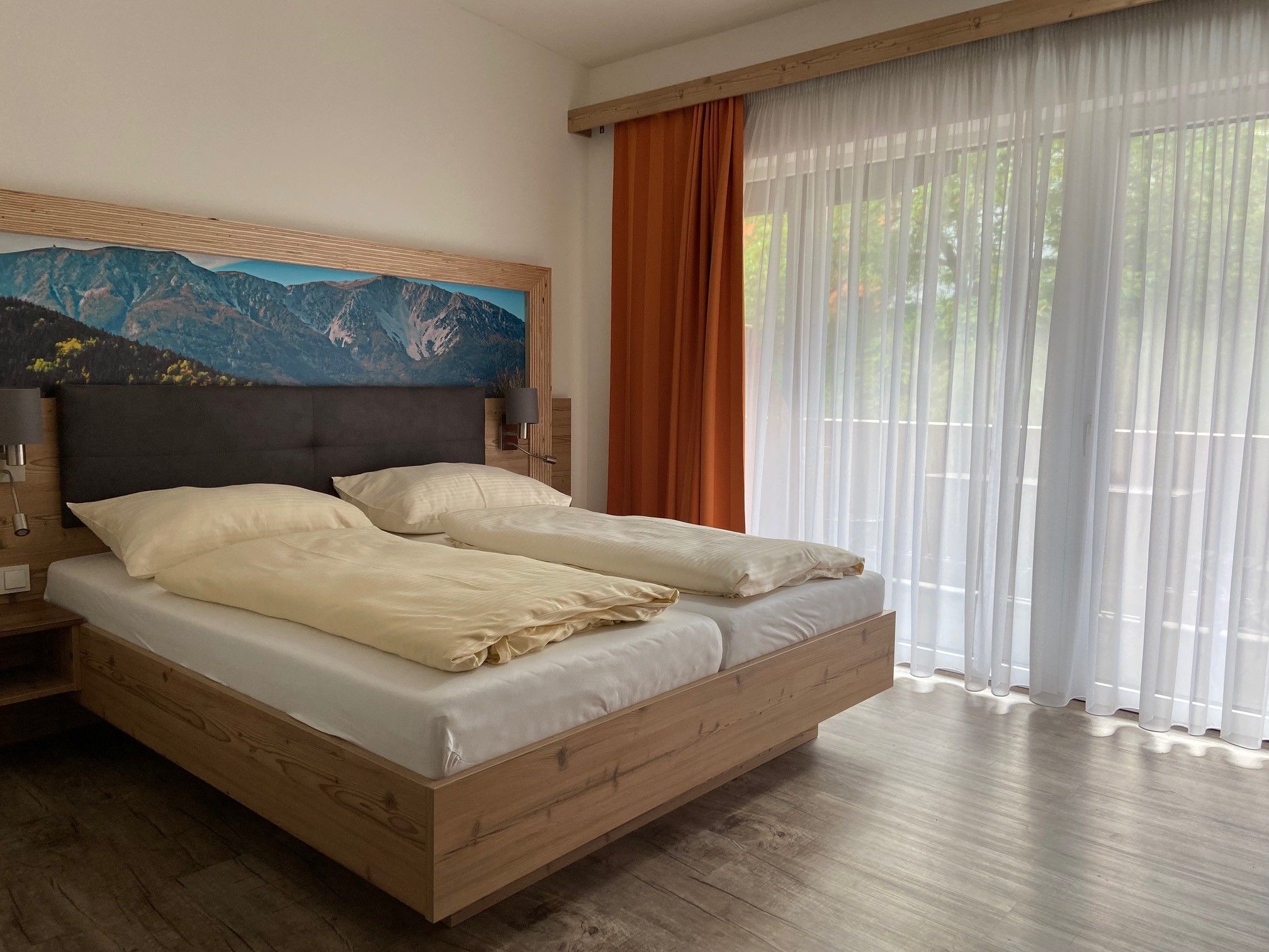 Modern double room with large bed, mountain picture on the wall and large windows with curtains.