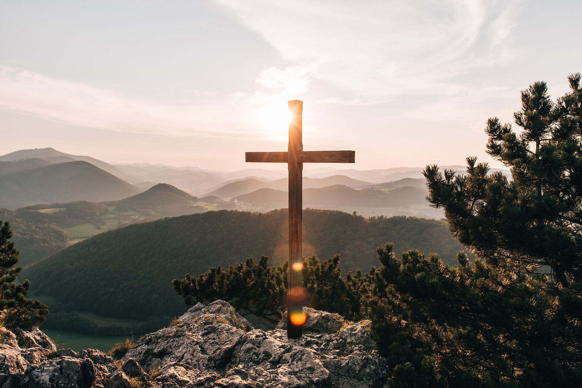 Ein majestätisches Kreuz erhebt sich vor der atemberaubenden Kulisse der sanften Hügel und Berge, während die Sonne langsam am Horizont aufgeht. Die goldenen Strahlen tauchen die Landschaft in ein warmes Licht und schaffen eine friedliche Atmosphäre, die zum Verweilen einlädt. Hier, inmitten der Natur, spürt man die Ruhe und Erhabenheit des Augenblicks.