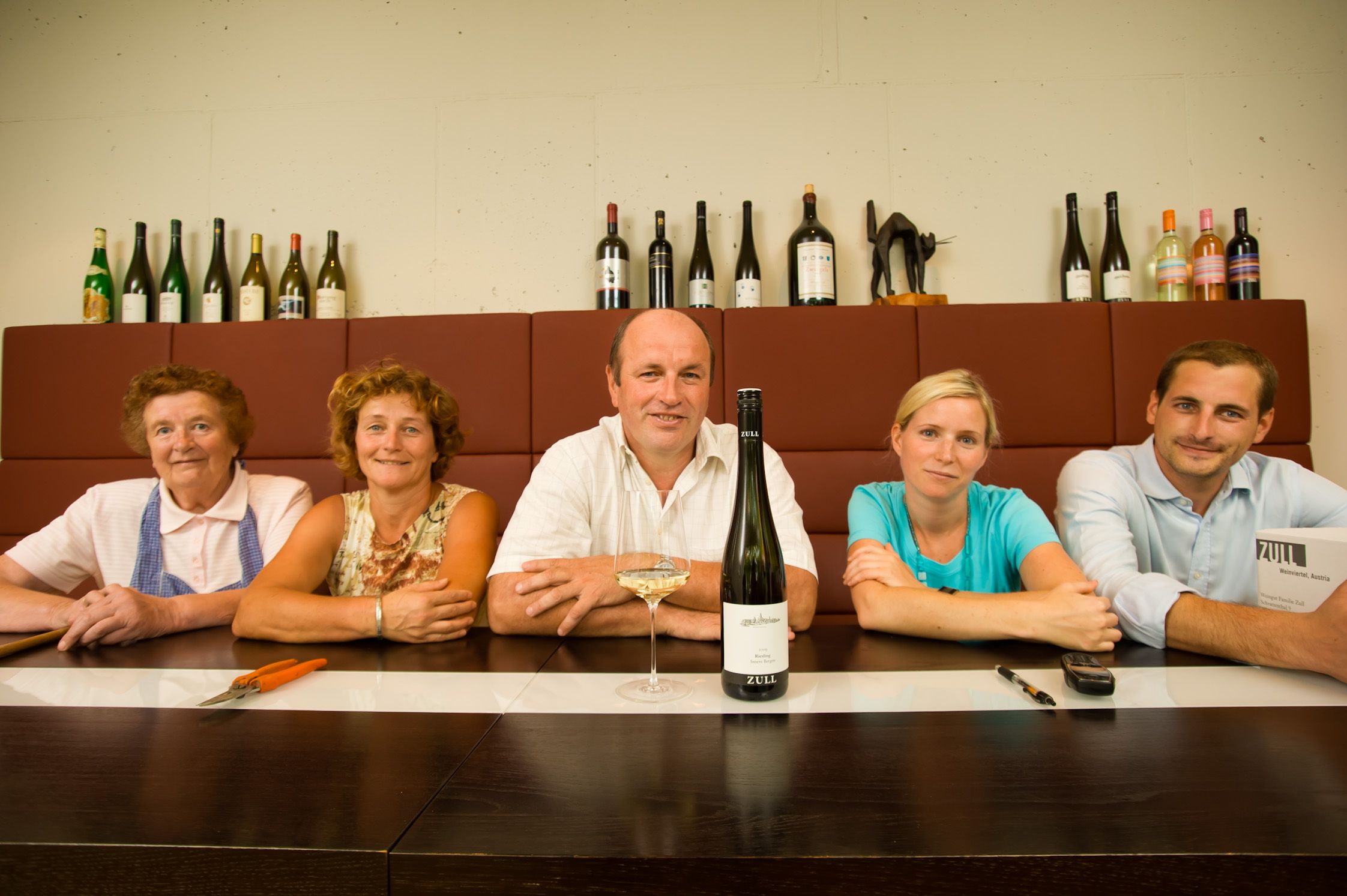 Five people sit at a table with bottles of wine in the background.