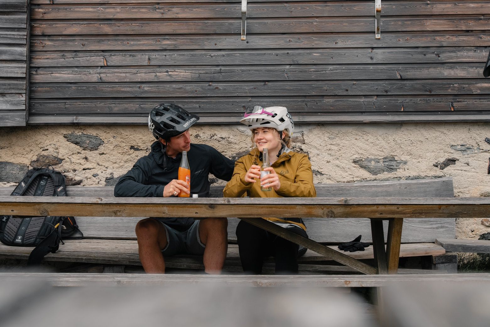 Two mountain bikers sit on a wooden bench in front of a hut and drink refreshments.