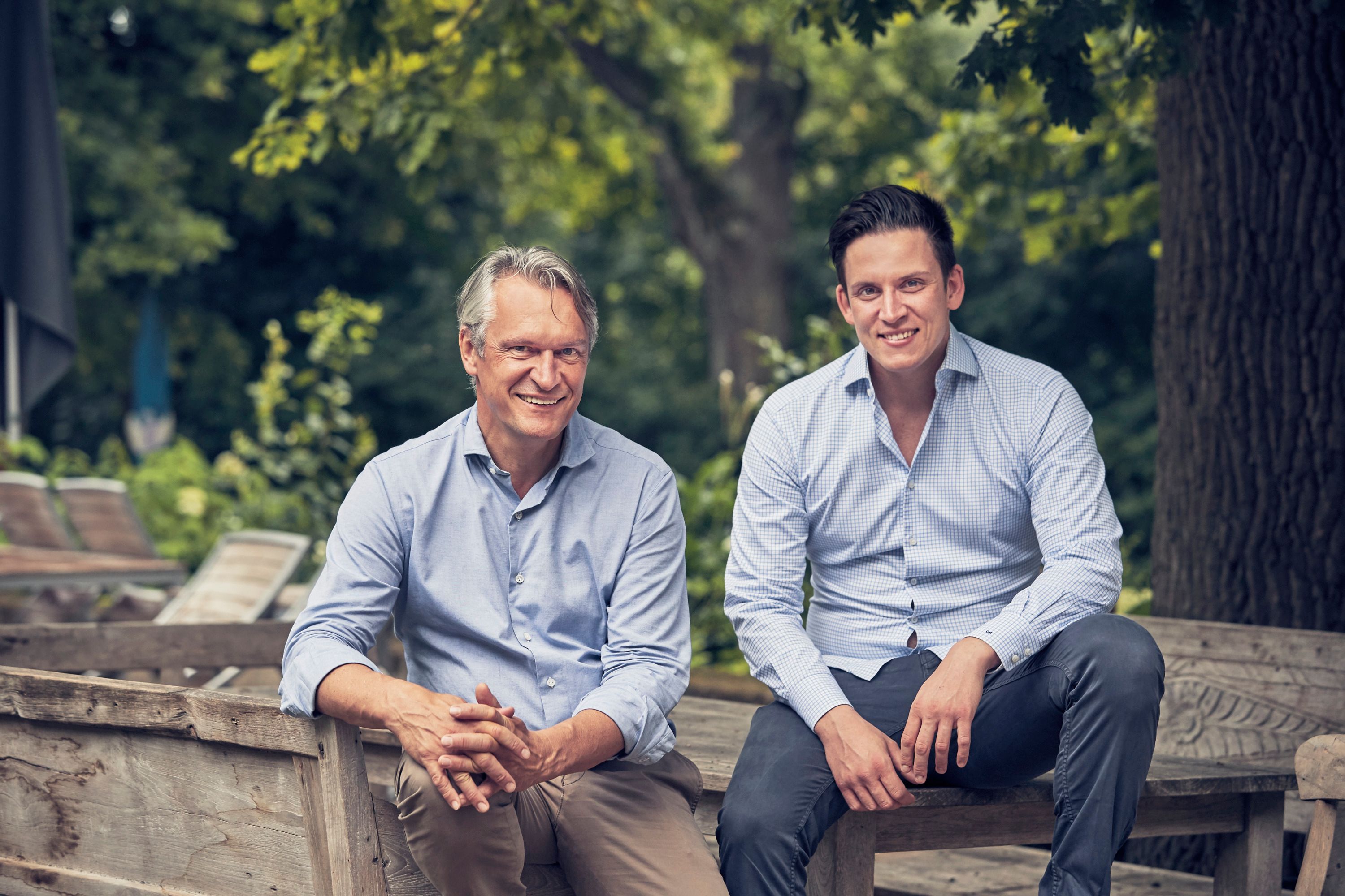 Two men are sitting on a wooden bench outside, surrounded by trees.