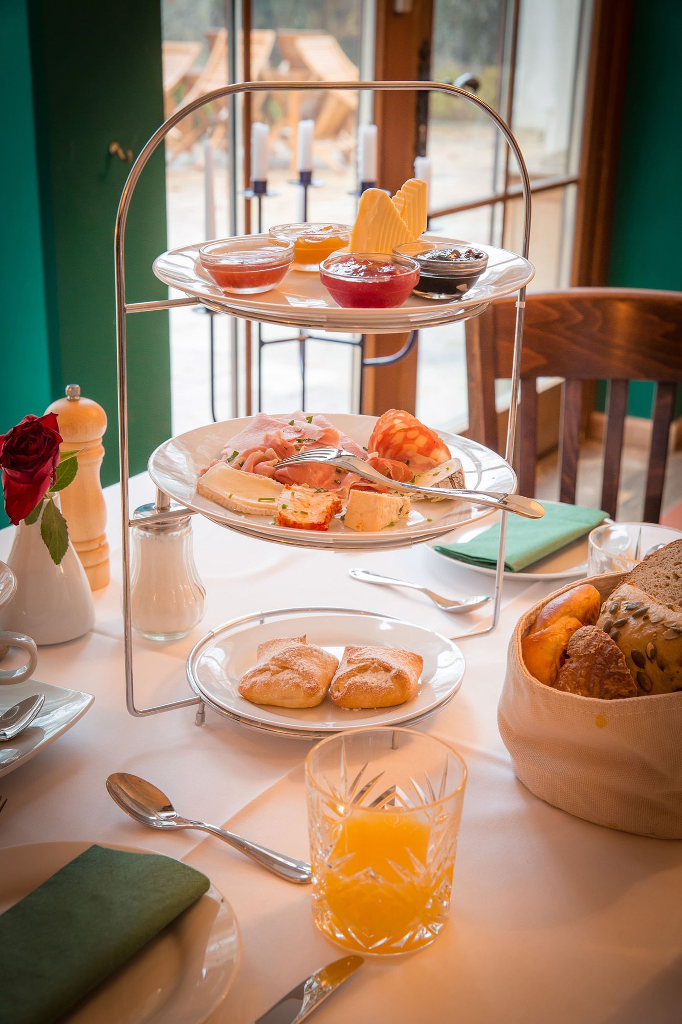 A breakfast table with an etagere full of cold cuts, jams and bread rolls, next to a glass of orange juice.