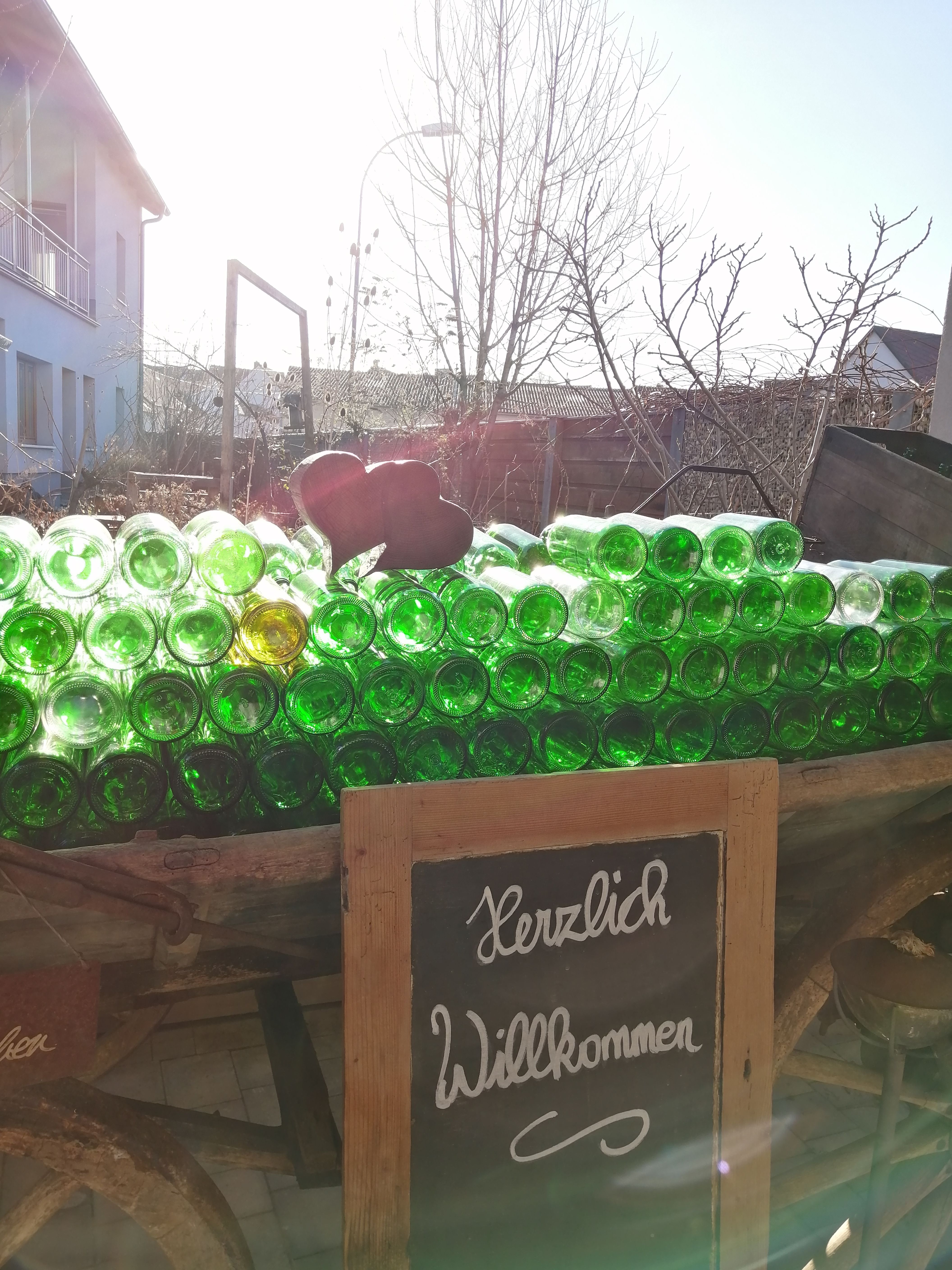 A pile of green glass bottles on a trolley with a 'Welcome' sign in front of it, a house and trees in the background.