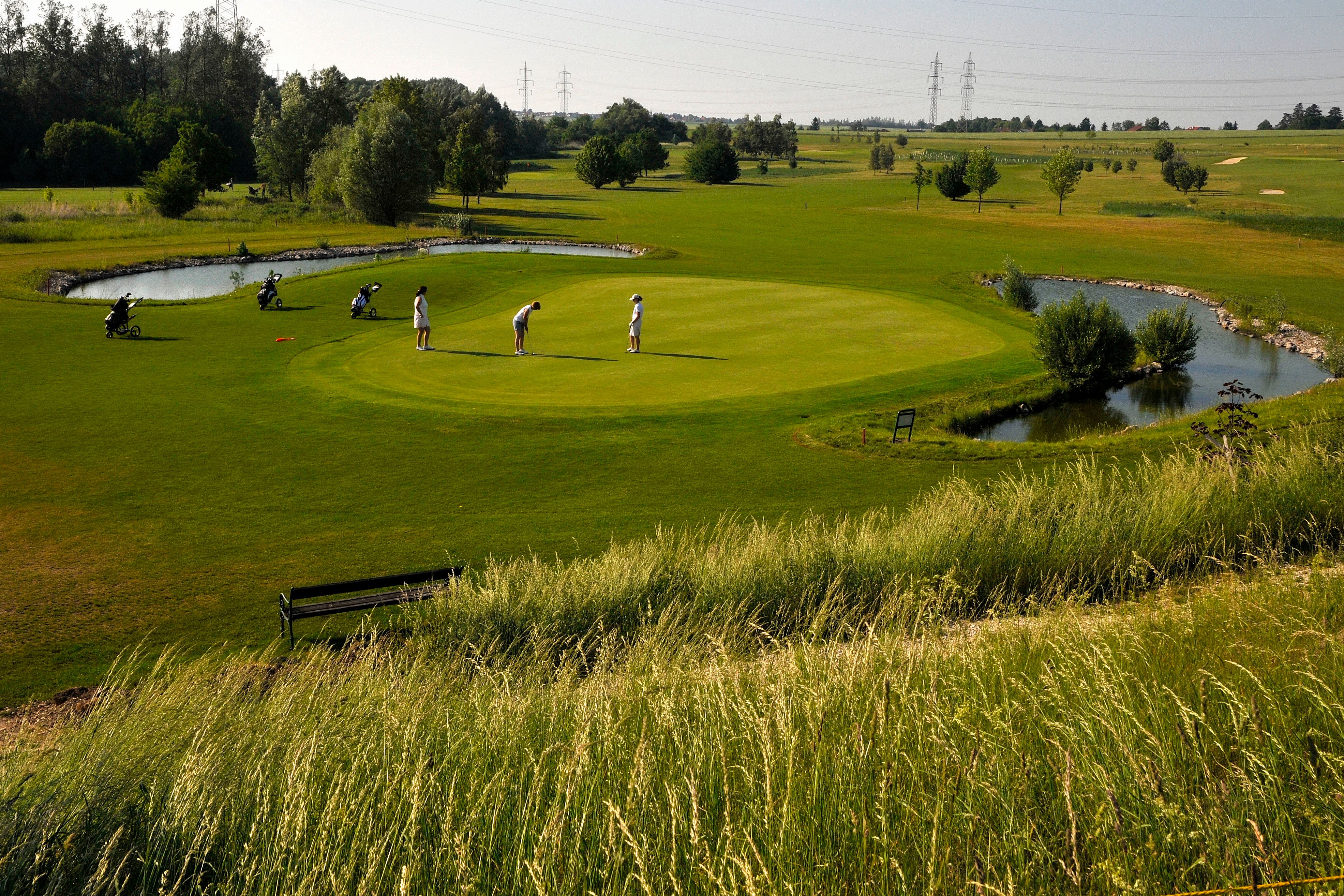 Golfers on a green golf course with a pond and trees in the background.