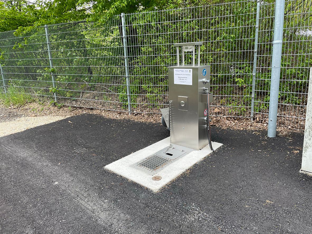 Metal water supply station next to a fence on asphalt.