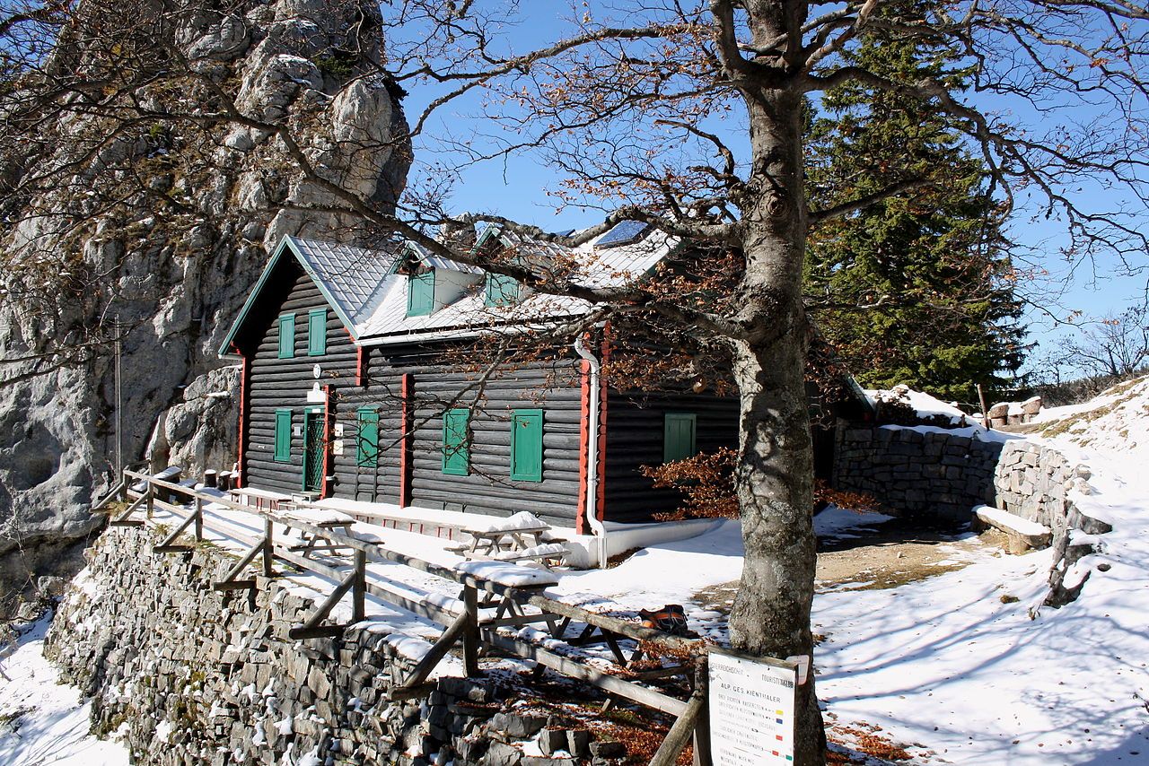 A wooden mountain hut with green shutters, surrounded by snow and rocks, under a clear blue sky.