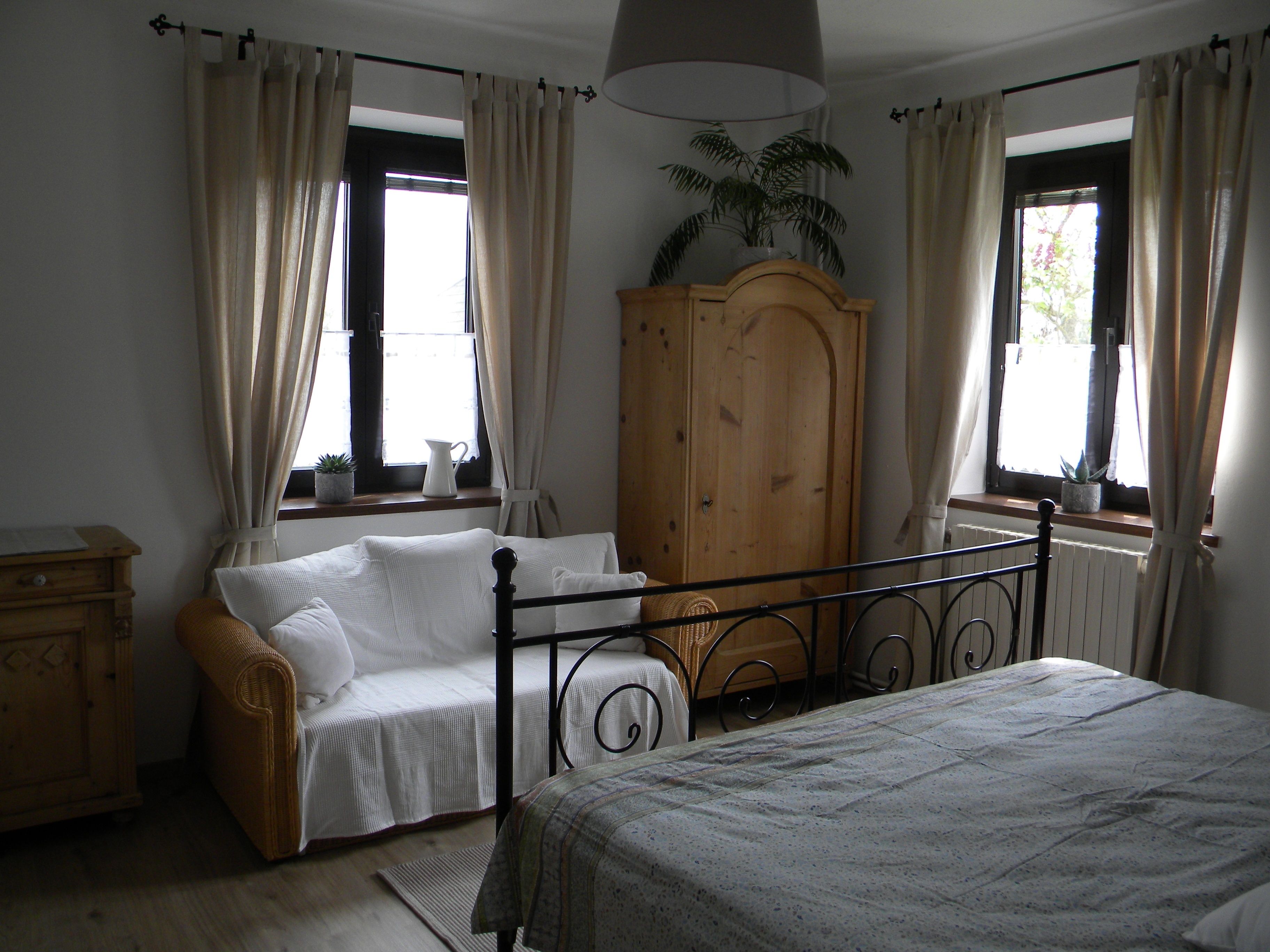 A cozy bedroom with a metal bed, a sofa, a wooden wardrobe and two windows with curtains.