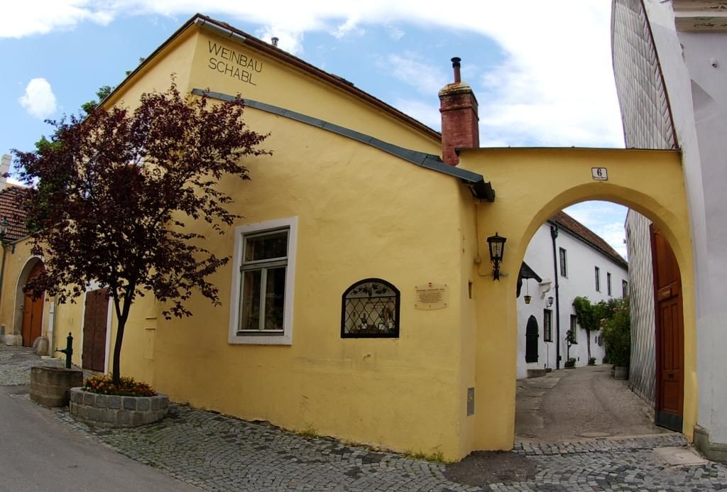 Yellow building with the inscription 'Weinbau Schabl', next to it an archway and a tree.