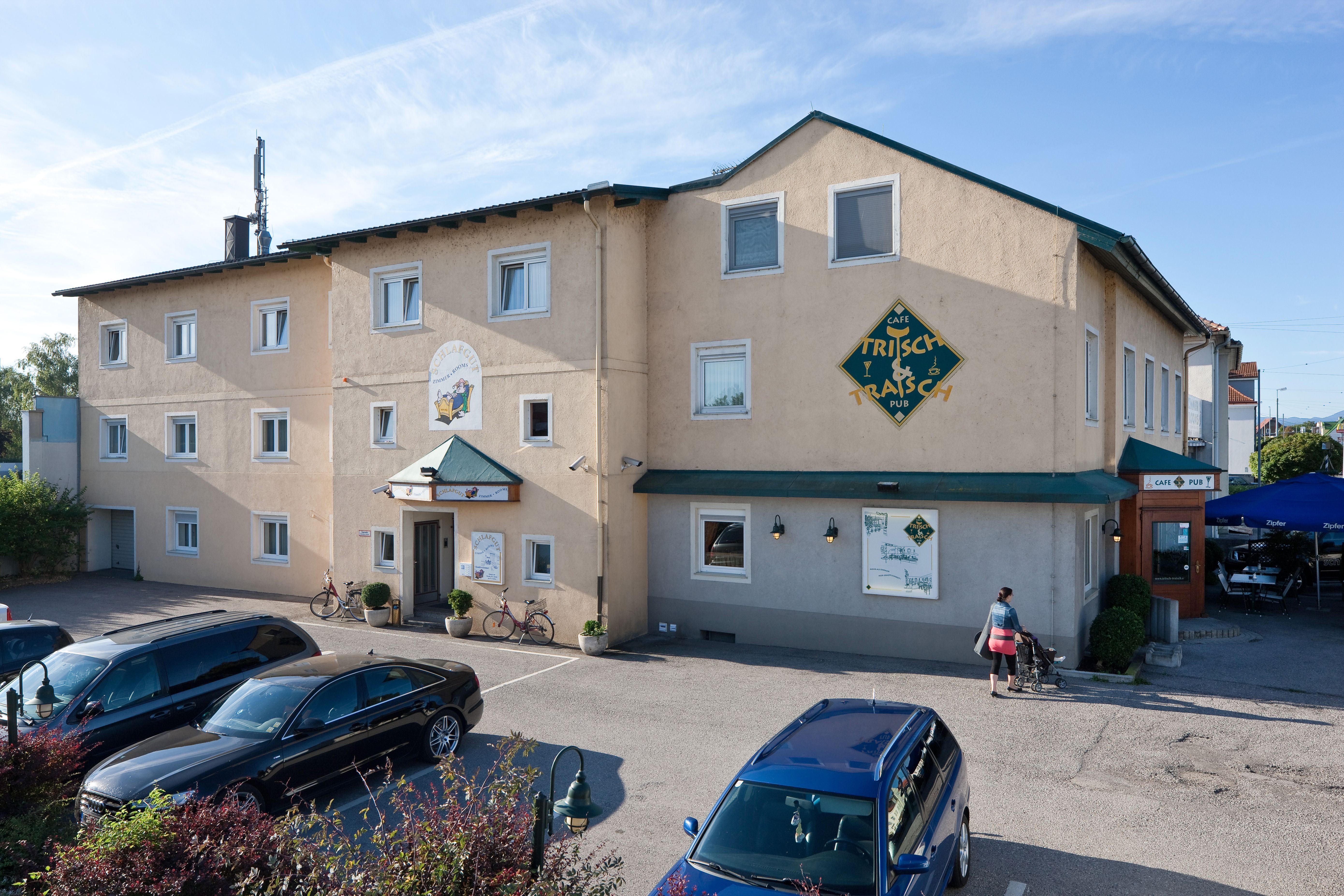 Exterior view of a building labeled 'Pension Schlafgut' and 'Café Trisch & Tratsch'. Cars are parked in front of it, a person with a baby carriage walks past.