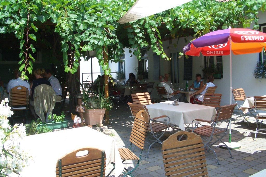 A cozy garden with tables and chairs under an arbor of vines, people sit and enjoy the atmosphere.