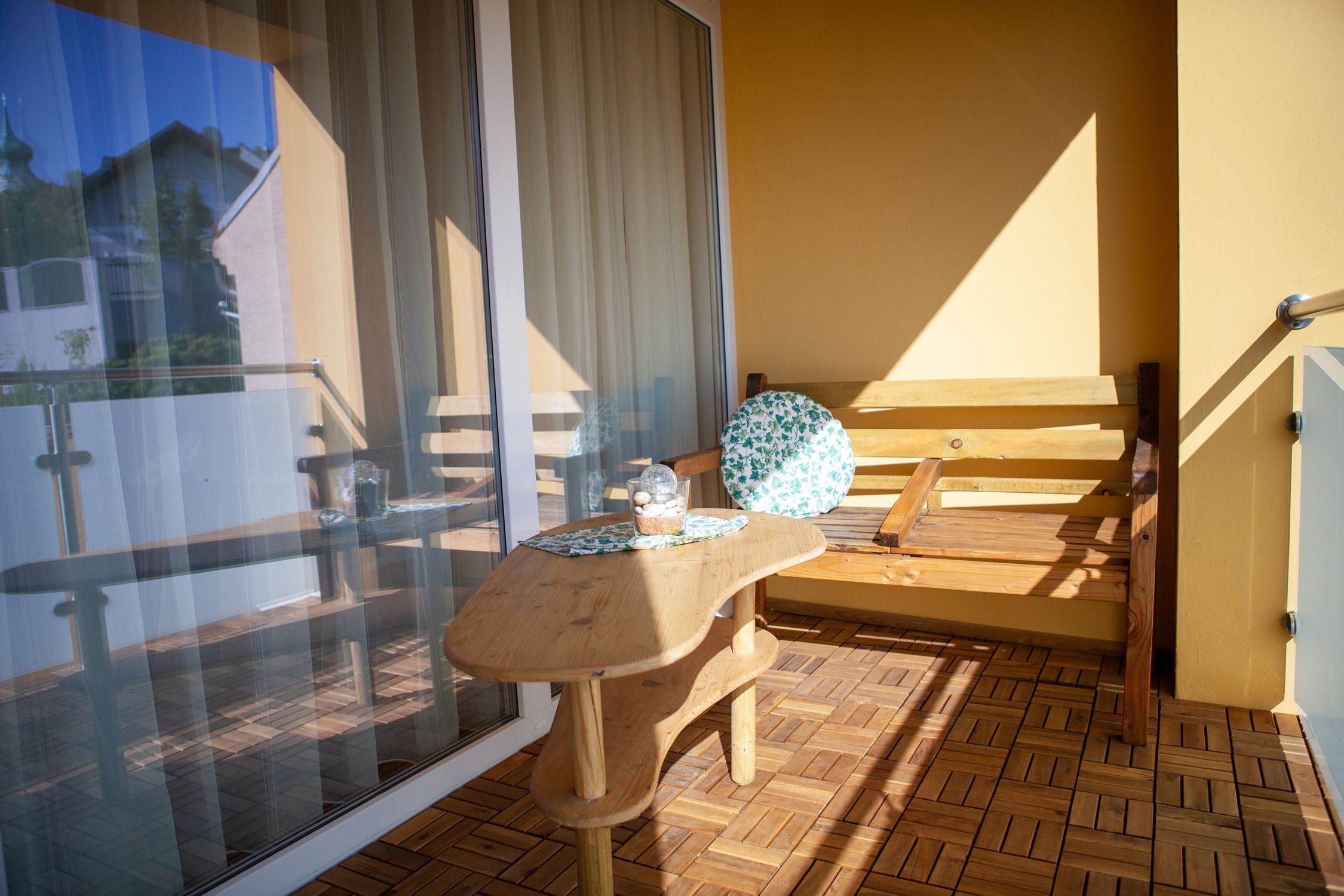 A balcony with a wooden floor, a wooden table and a wooden bench with a round cushion. A glass railing and a building can be seen in the background.