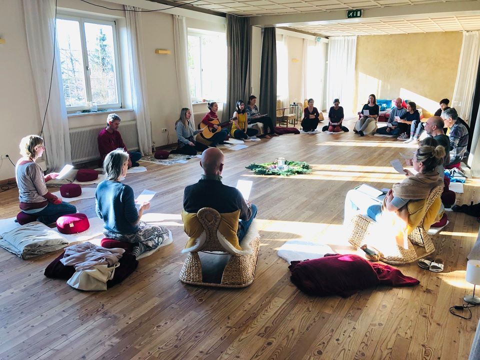 People sit in a circle on cushions and chairs in a bright seminar room with a wooden floor.