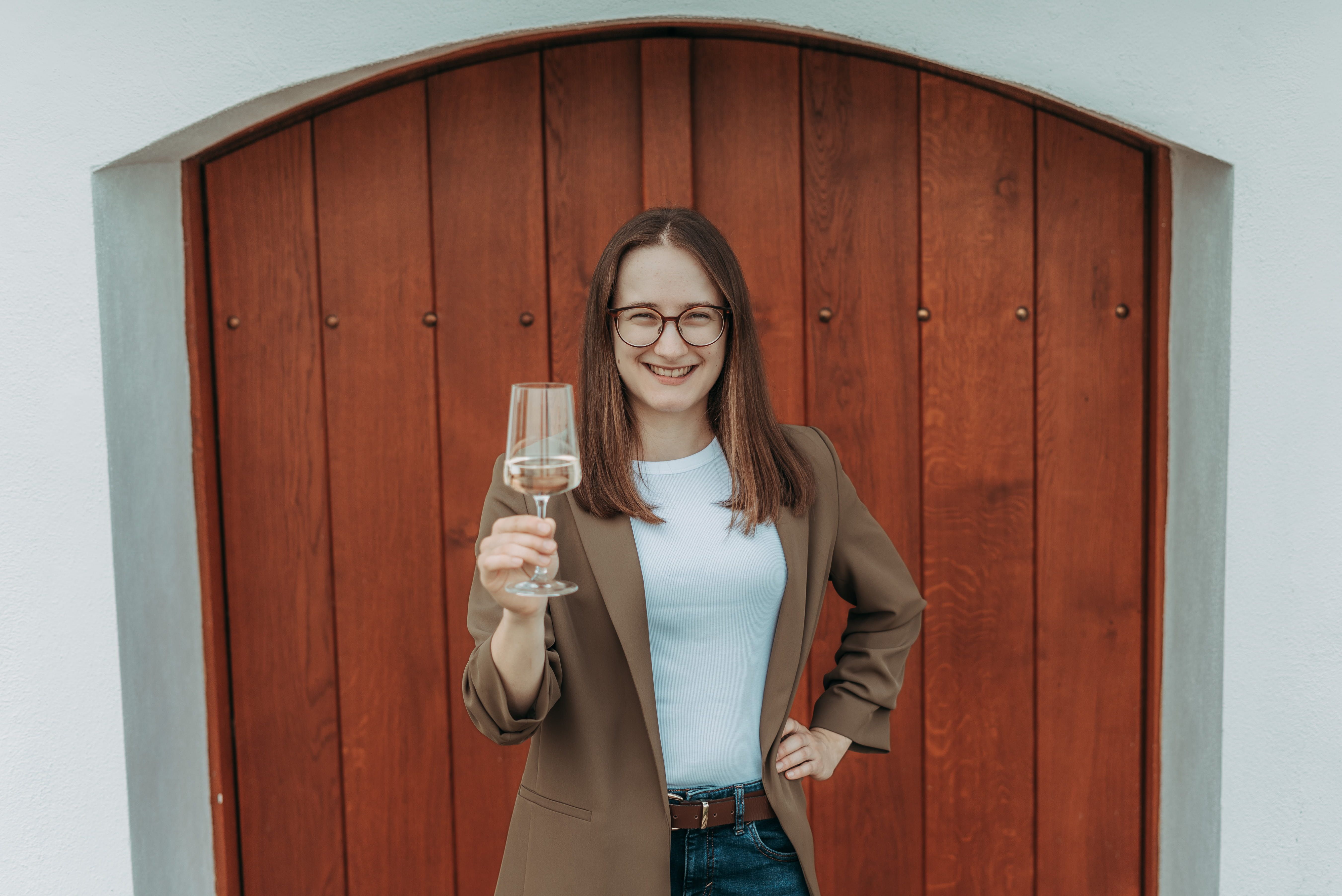 A woman with glasses holds a wine glass and smiles in front of a wooden door.