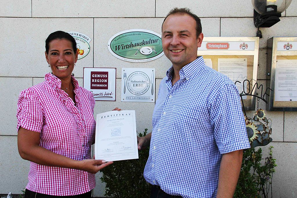 Two people holding a certificate in front of a wall of awards.