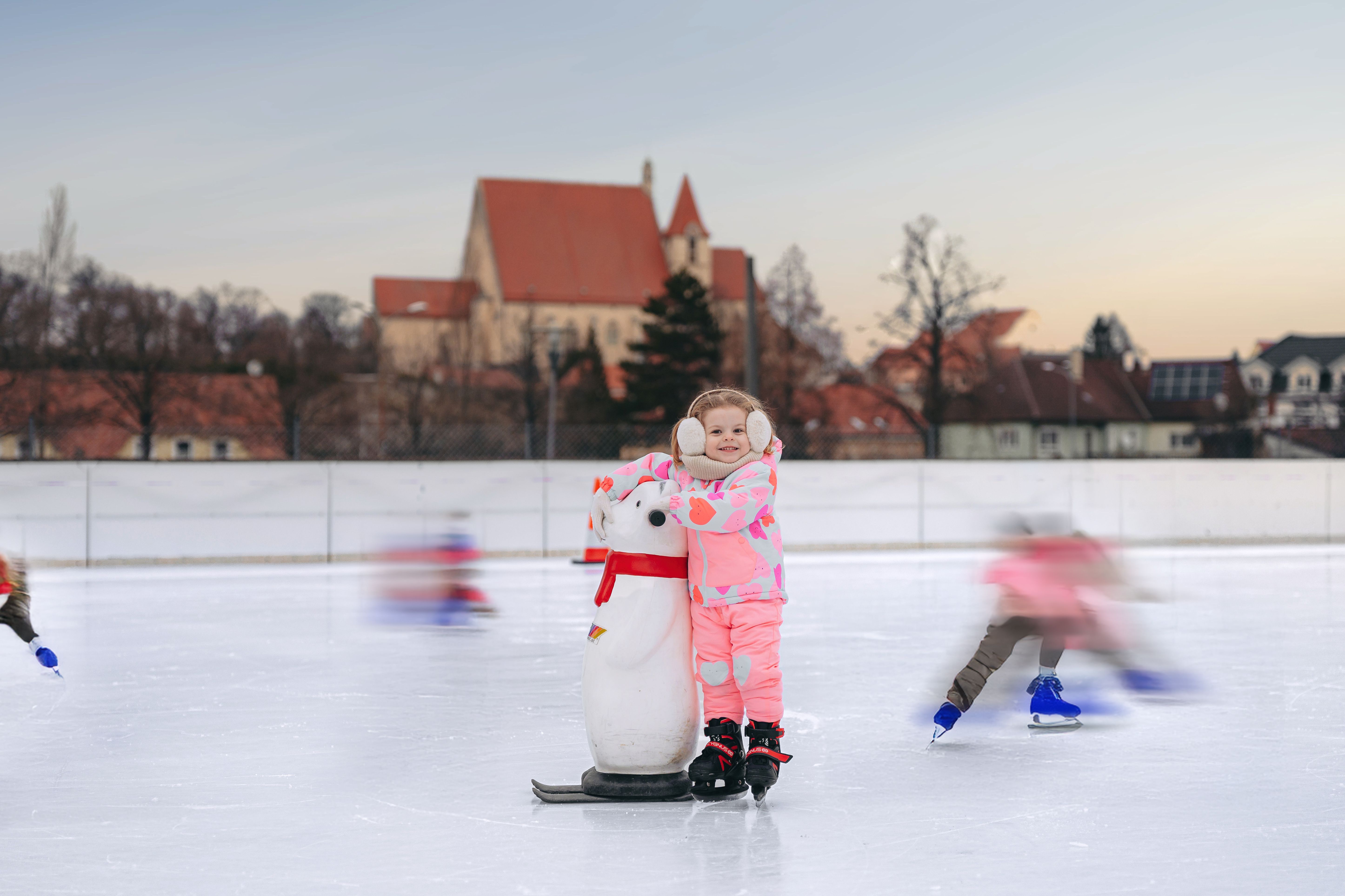 A child in pink clothing stands on an ice rink with a penguin aid. A church can be seen in the background.