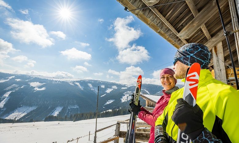 Two people on skis stand in front of a wooden hut in a snowy mountain landscape in the sunshine.
