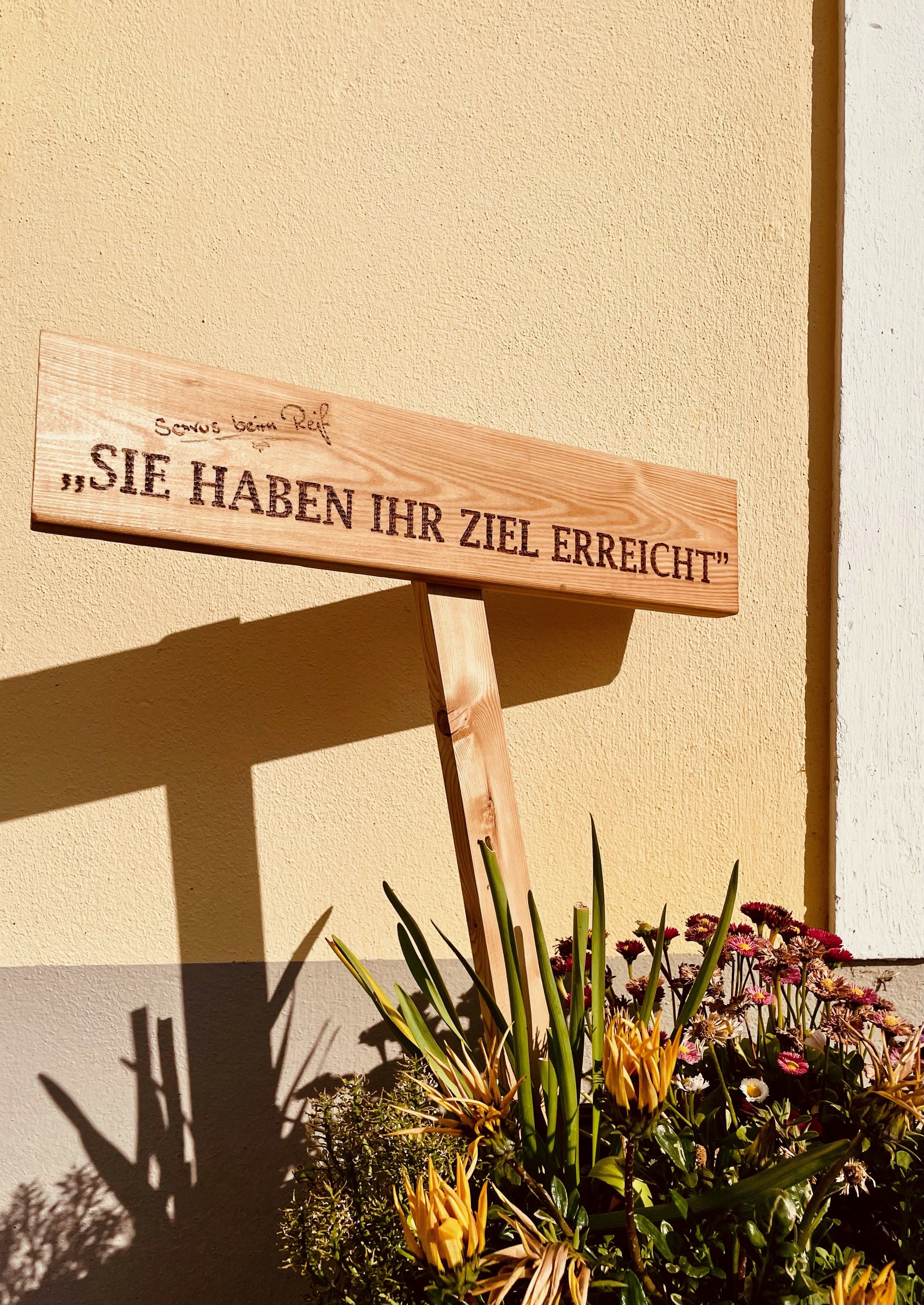 Wooden sign with the inscription 'You have reached your destination' in front of a yellow wall surrounded by flowers.