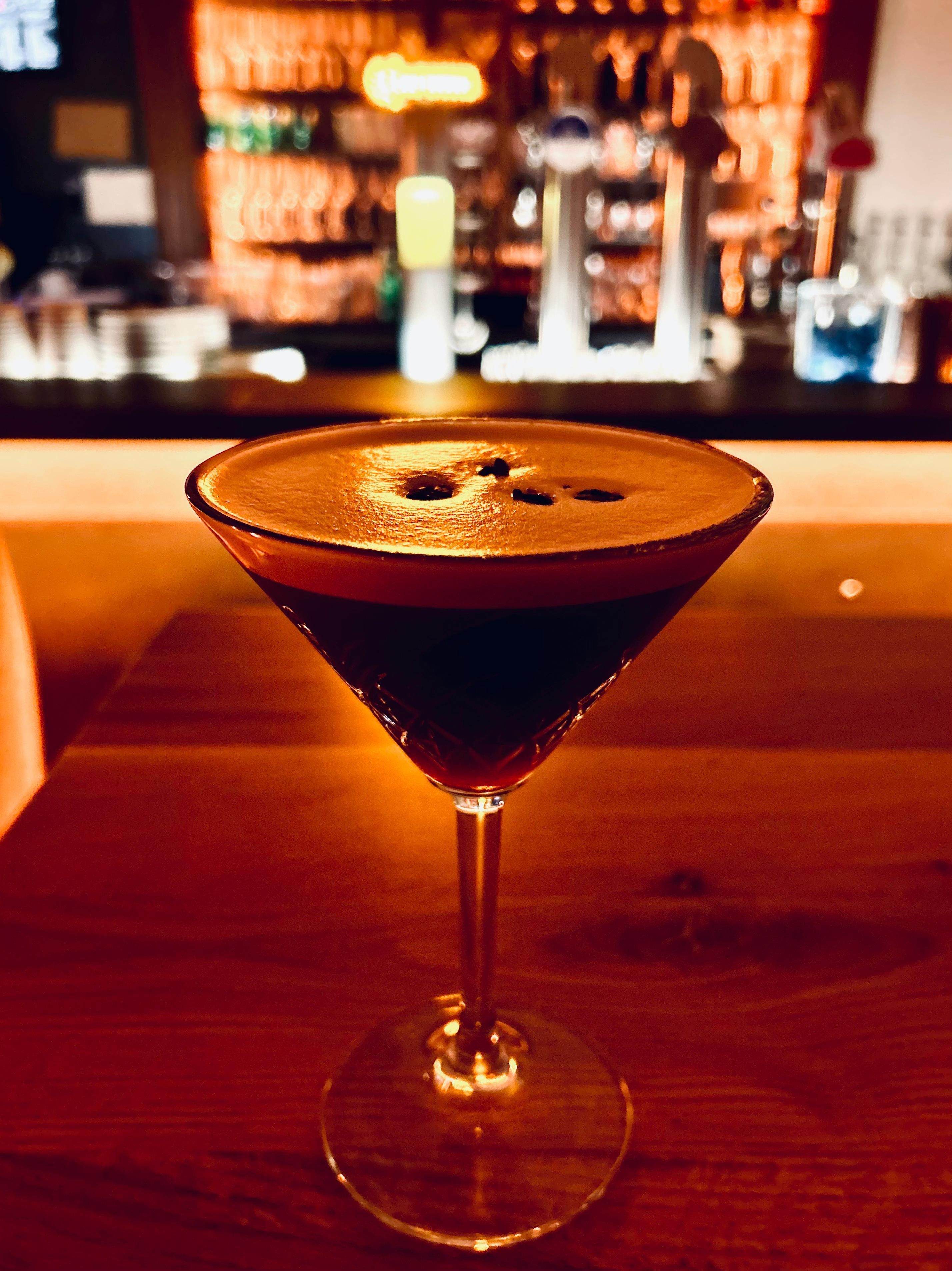A cocktail glass with a dark drink on a wooden table, a blurred bar in the background.