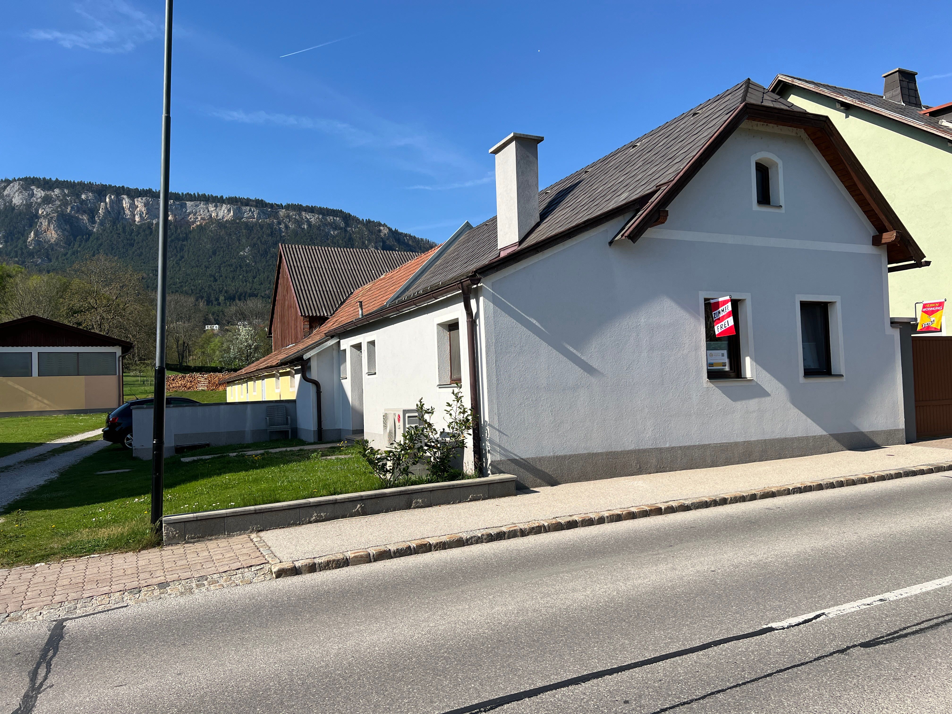 A white house with a red roof and a "For Rent" sign in front of a mountain backdrop.