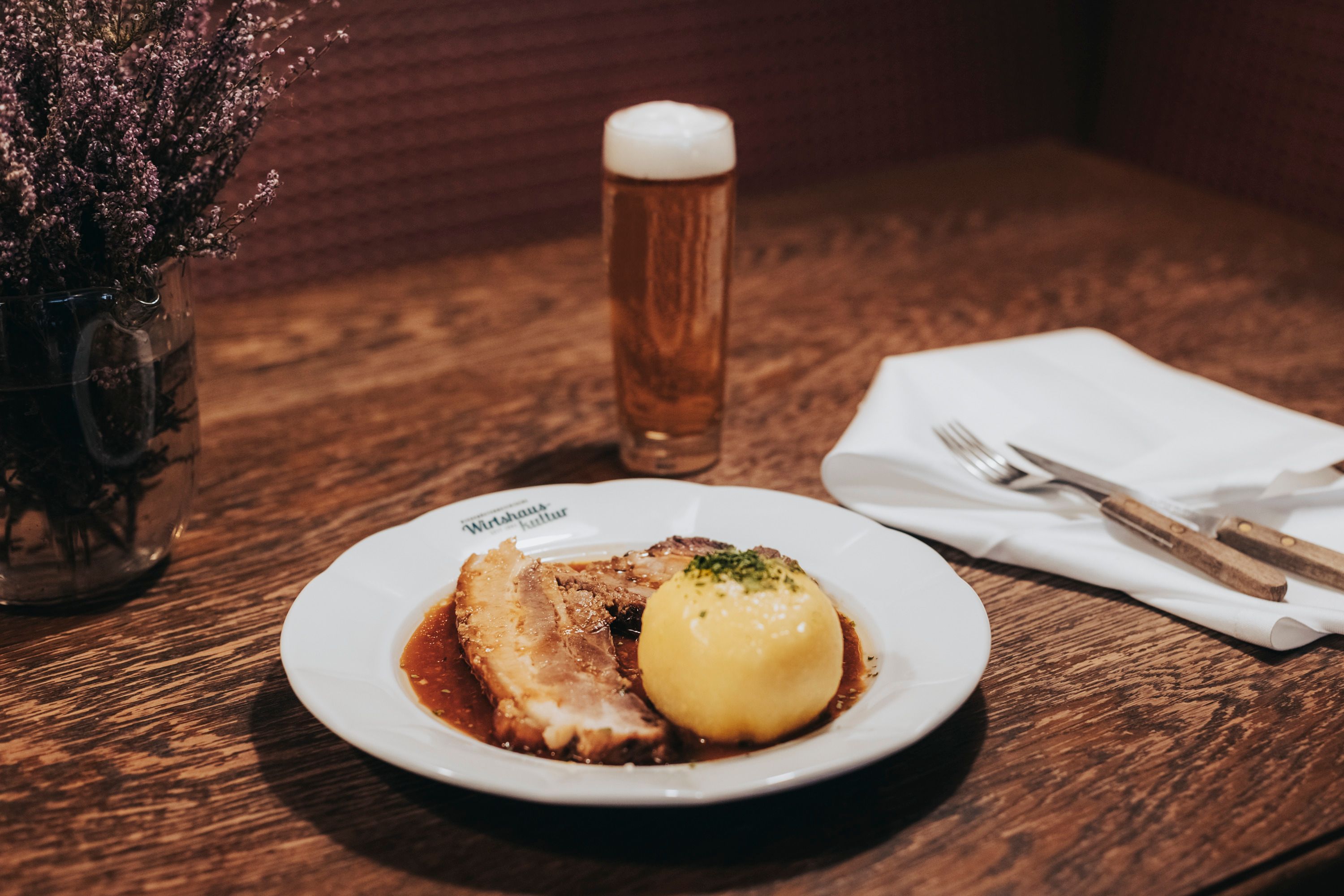 A plate of roast pork and Waldviertler dumplings, next to a glass of beer on a wooden table.