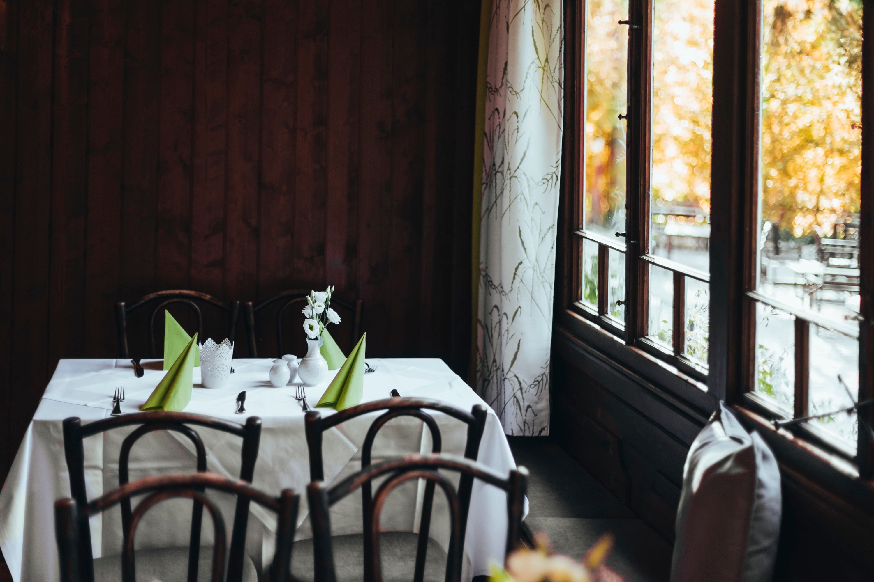 A table in an inn with wooden chairs, white tablecloths and green napkins, next to a window with curtains.