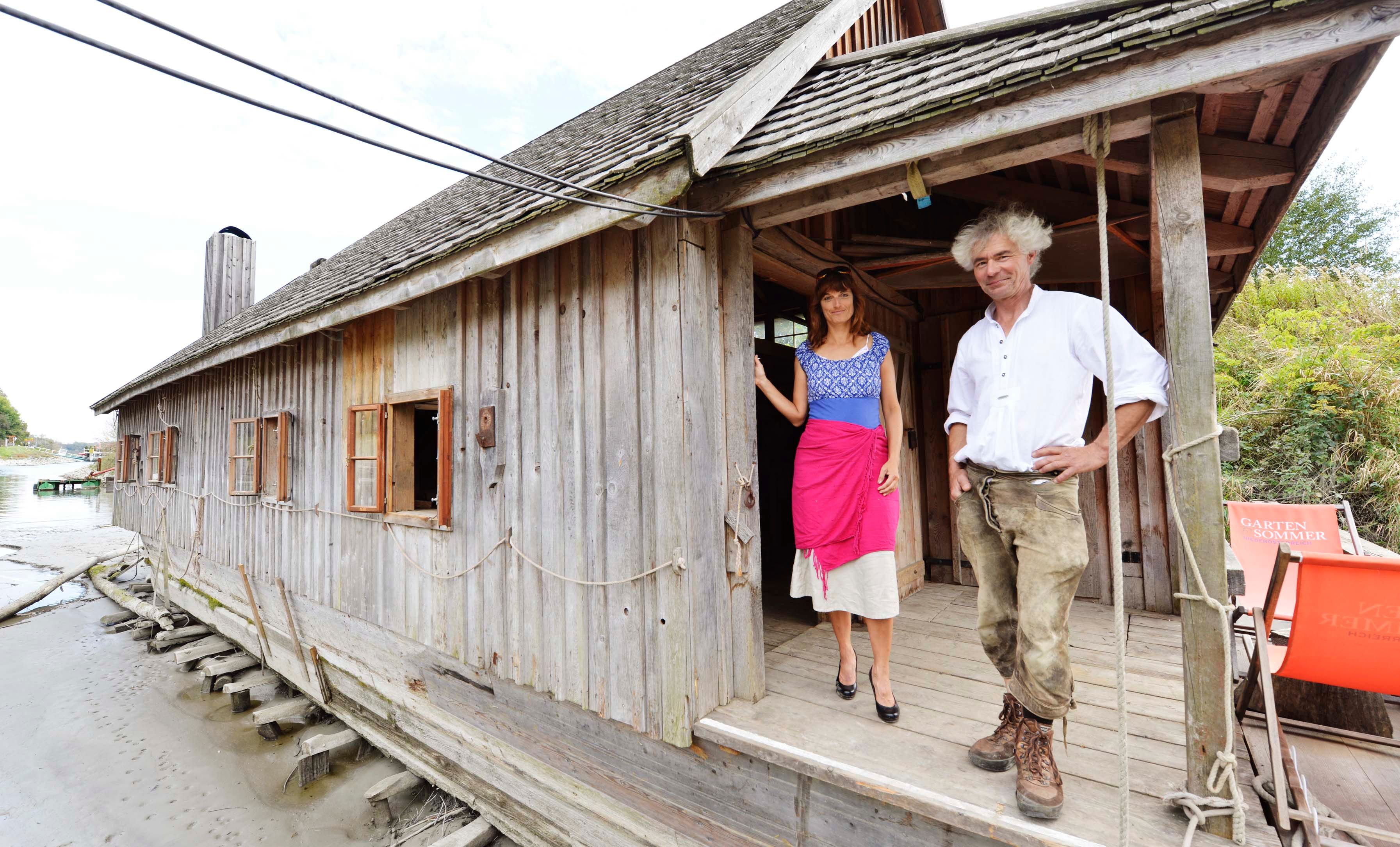 Two people are standing on the veranda of a wooden ship's mill on a riverbank.