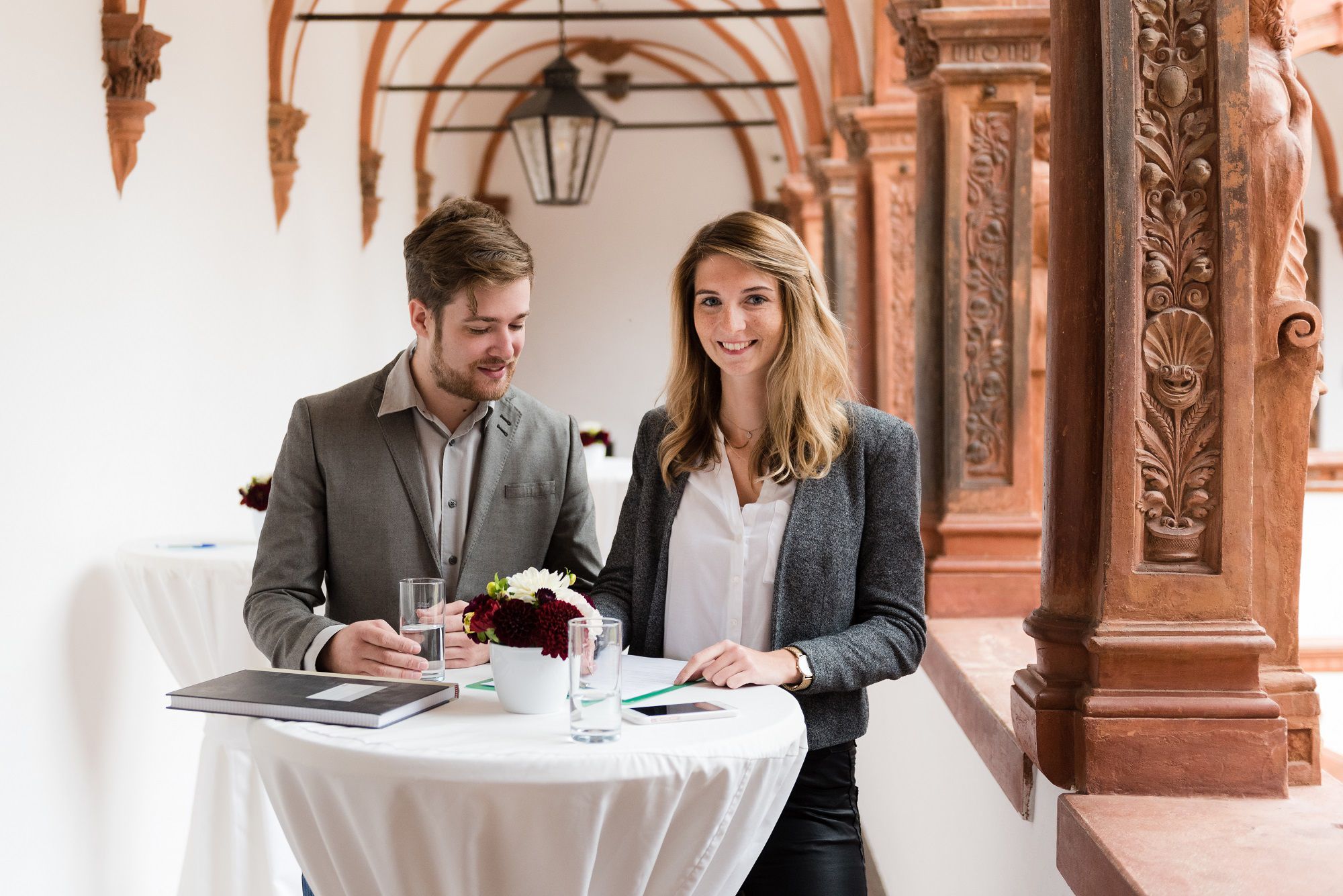 Two people are standing at a high table in a historic building with ornate columns.