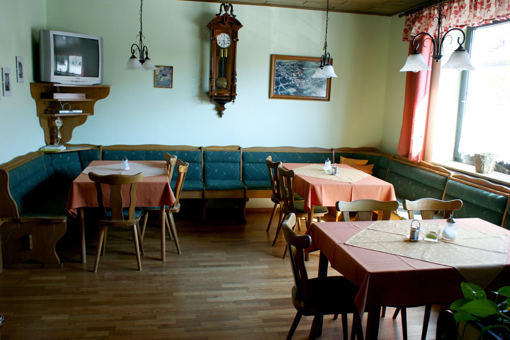 A traditionally furnished breakfast room with wooden furniture, tables with orange tablecloths, a wall clock and a television.