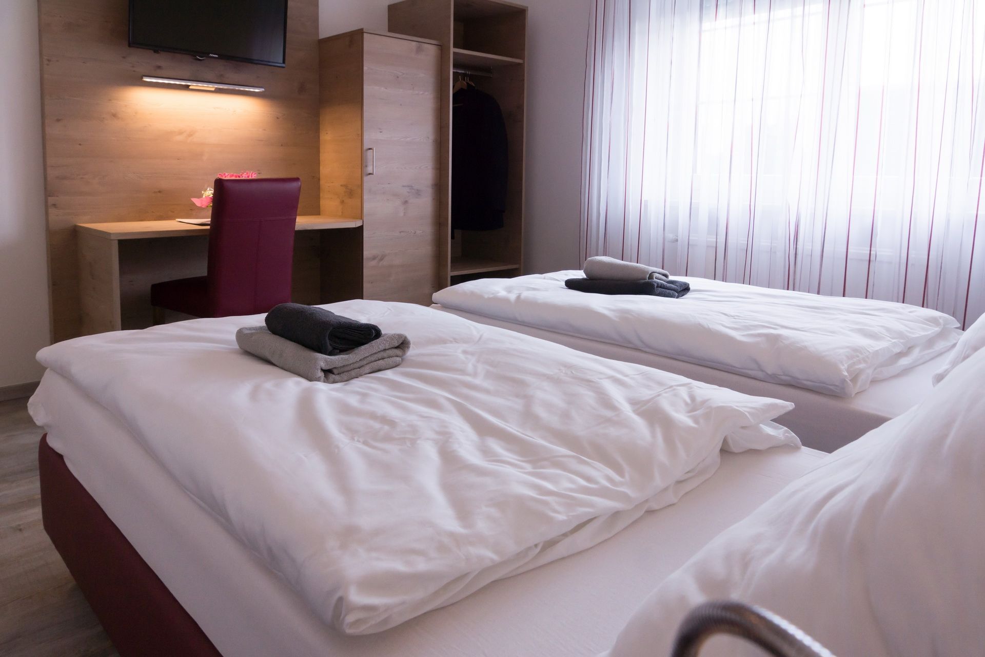 Twin room with two beds, desk, chair and closet. Towels on the beds, window with curtains in the background.