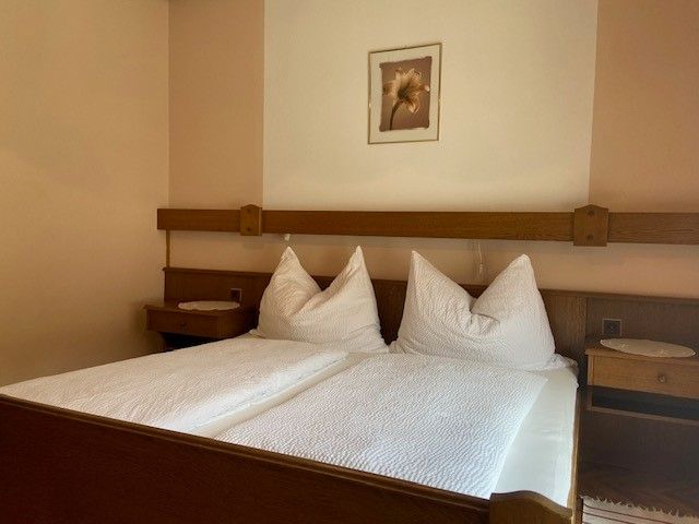 A cozy double room with a large bed, white pillows and blankets, wooden furniture and a flower picture on the wall.