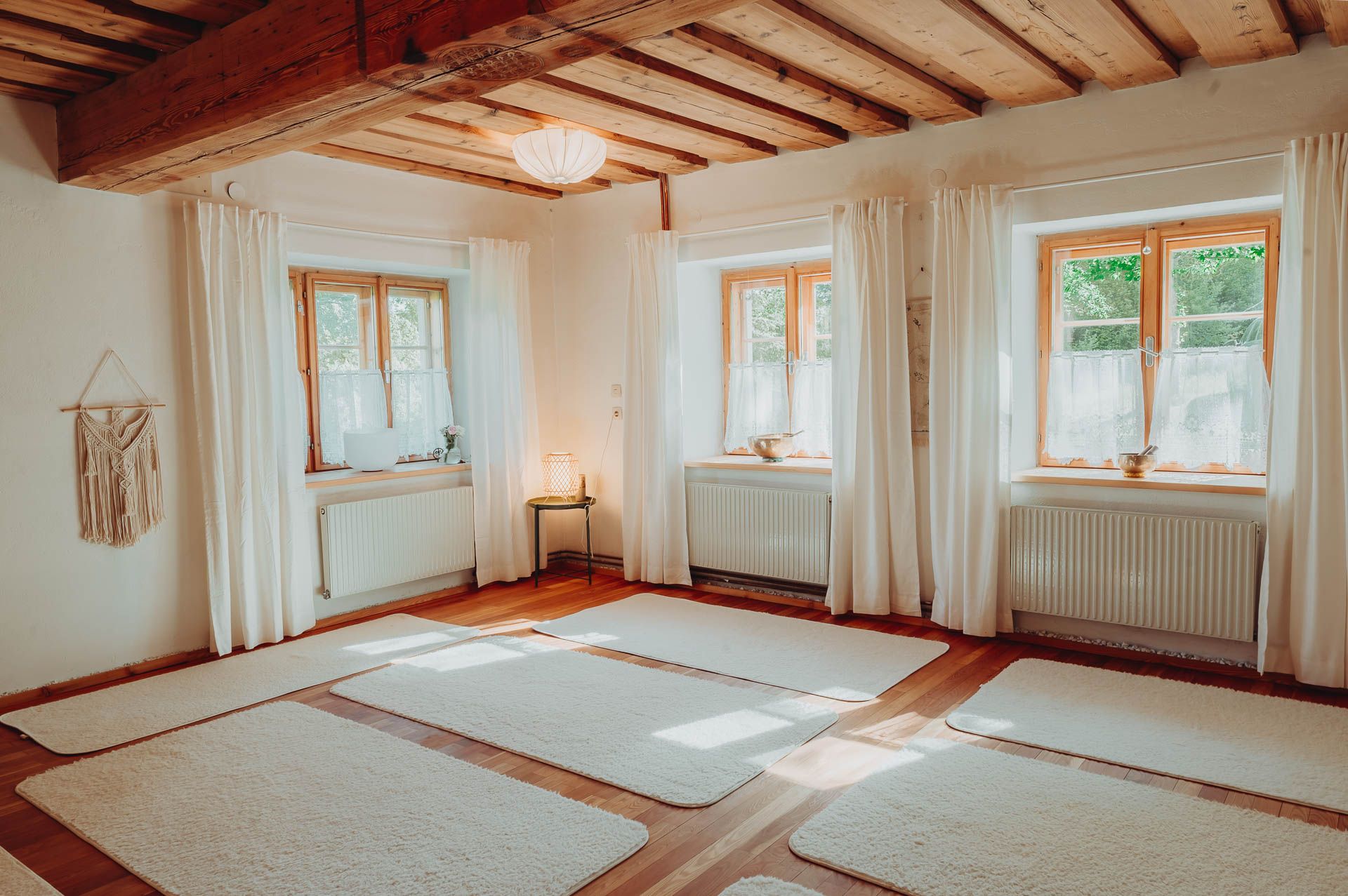 A bright yoga room with a wooden floor, white mats and large windows with curtains.