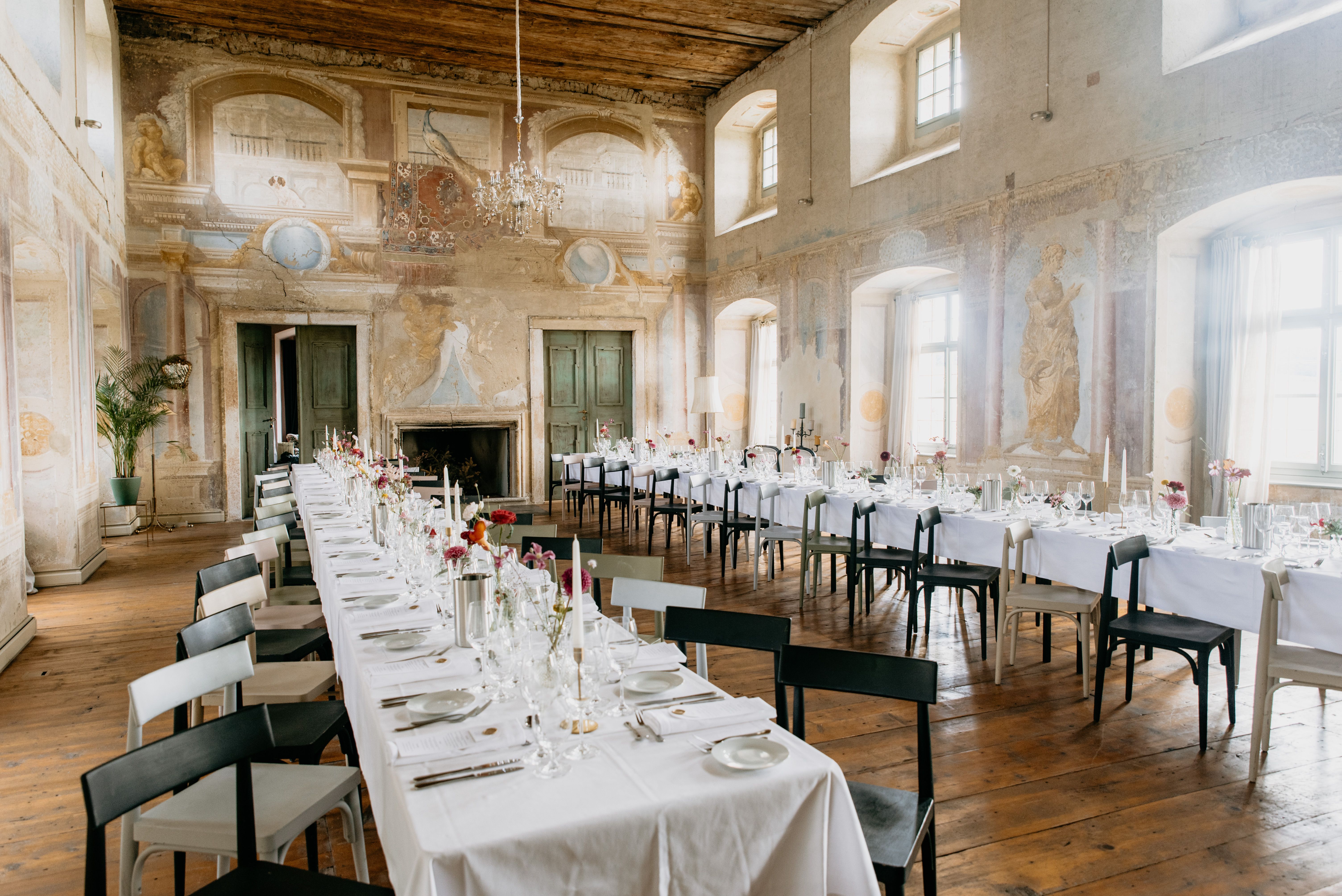 Elegant ballroom with long, laid tables, antique wall paintings and chandeliers.
