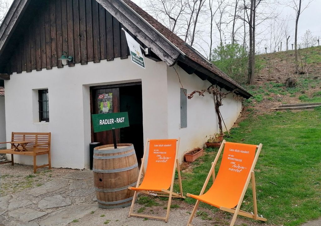 A small white building with a wooden roof, two orange deckchairs and a wooden barrel in front of it, surrounded by trees and grass.