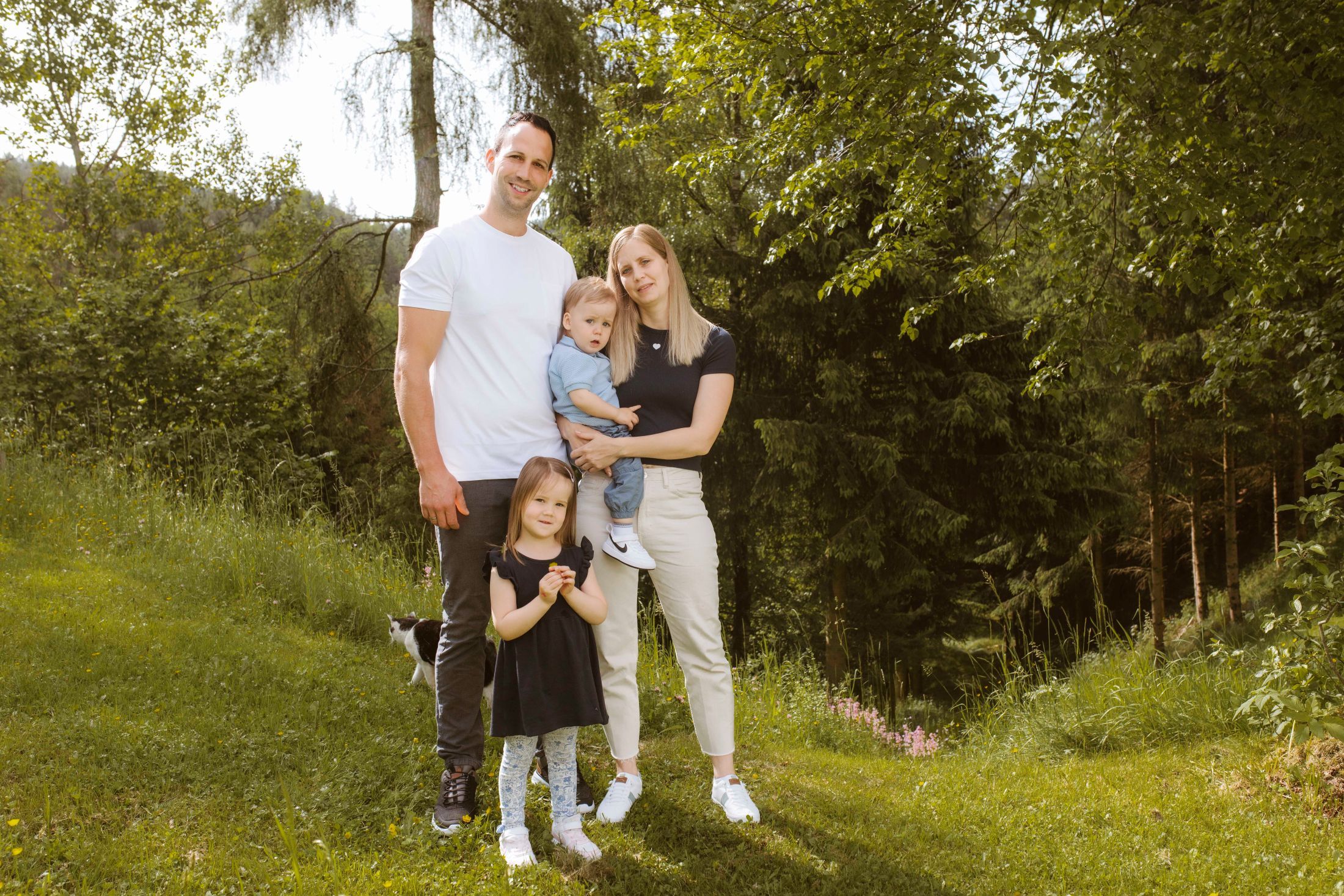 A family is standing in a meadow in front of a forest. A man, a woman, a small child in the woman's arms and another child are standing together.