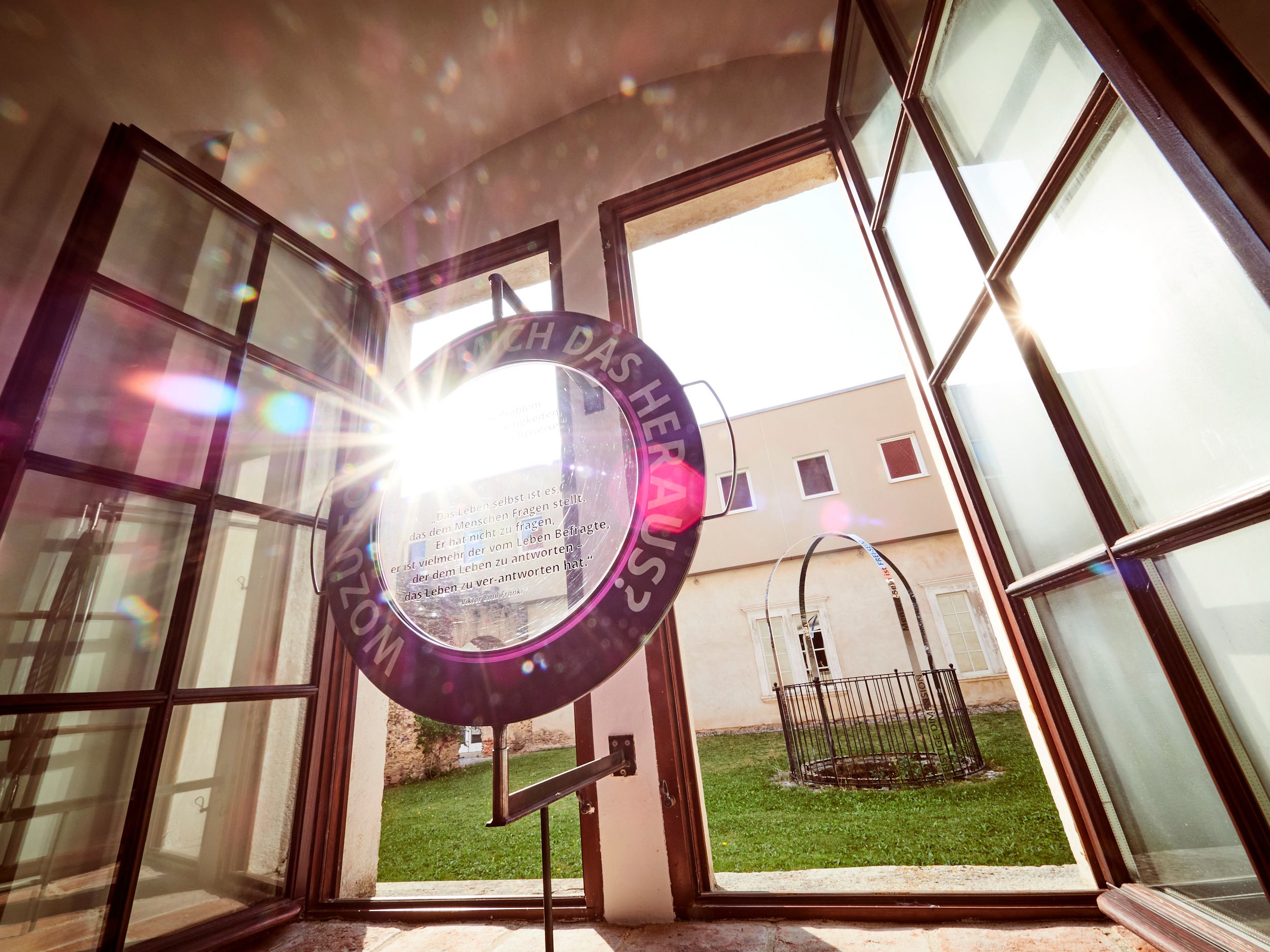 An open window with a round work of art through which sunlight shines and a view of a green inner courtyard.
