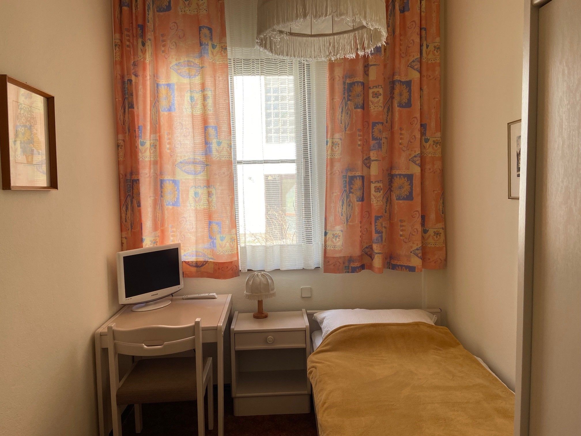 A small single room with a bed, desk, chair and television. Colorful curtains and a lamp can also be seen.