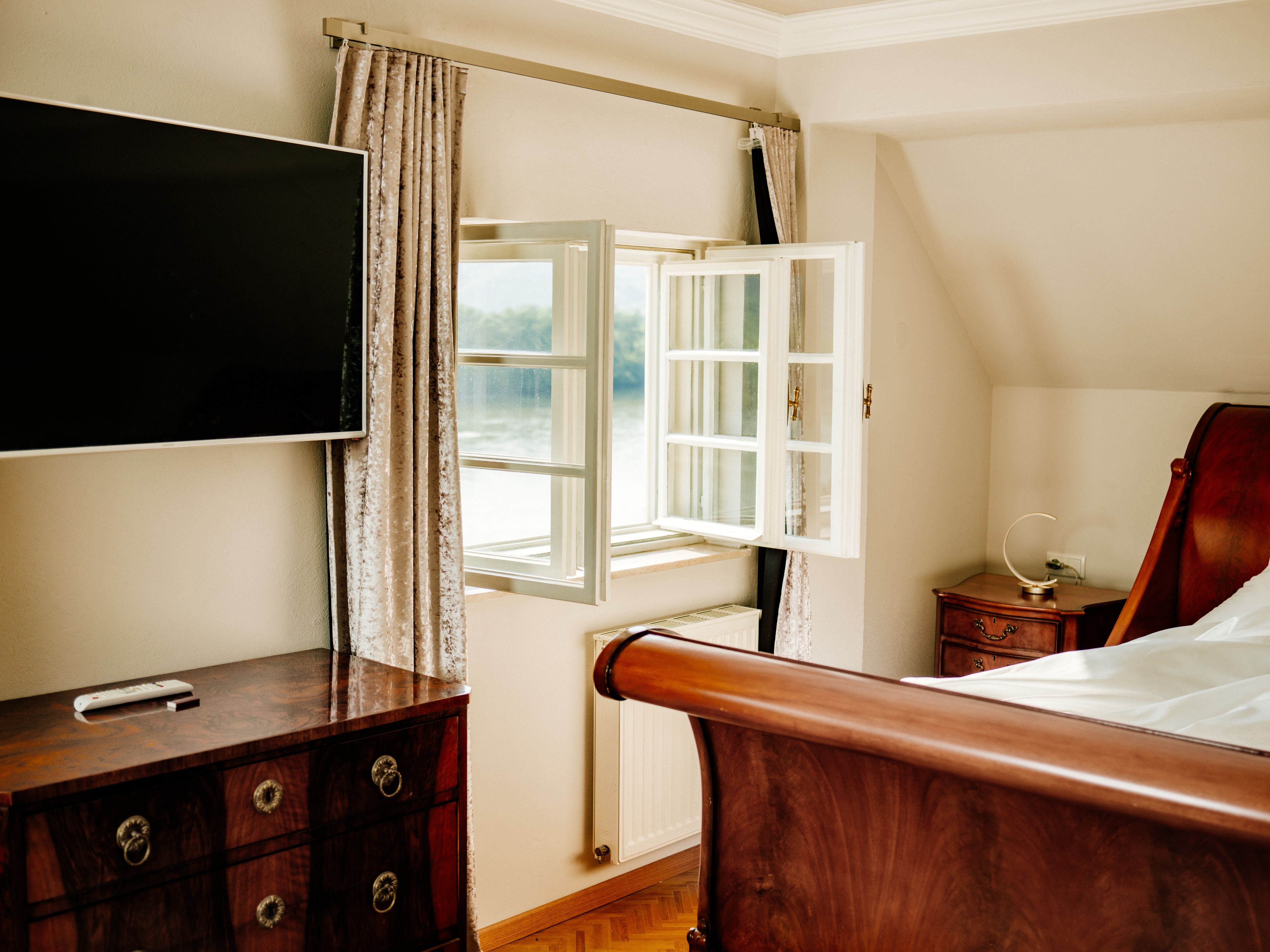 An elegant bedroom with an open window, a flat-screen TV, a wooden bed and antique furniture.