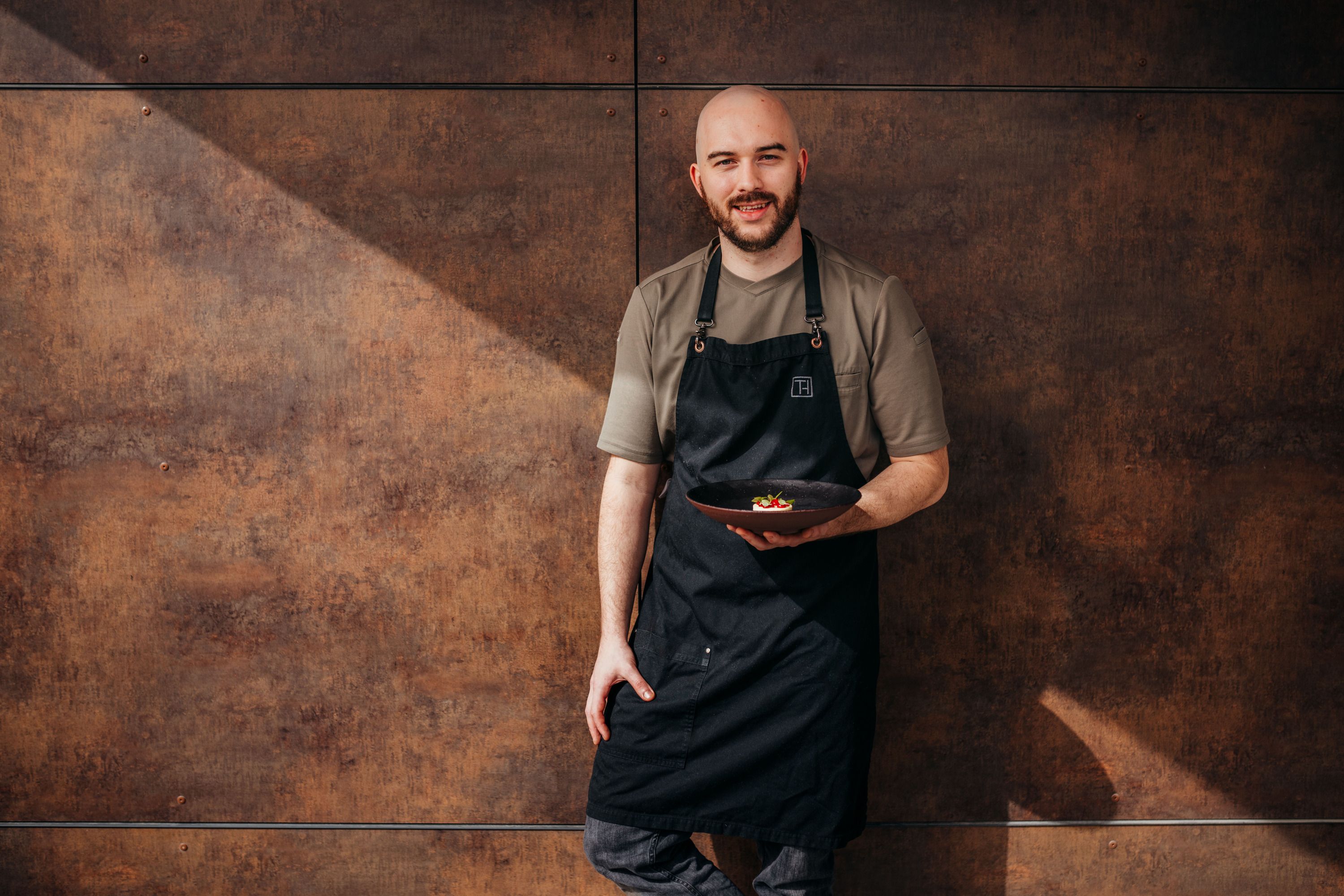 A man in a black apron holds a plate of food in front of a brown wall.