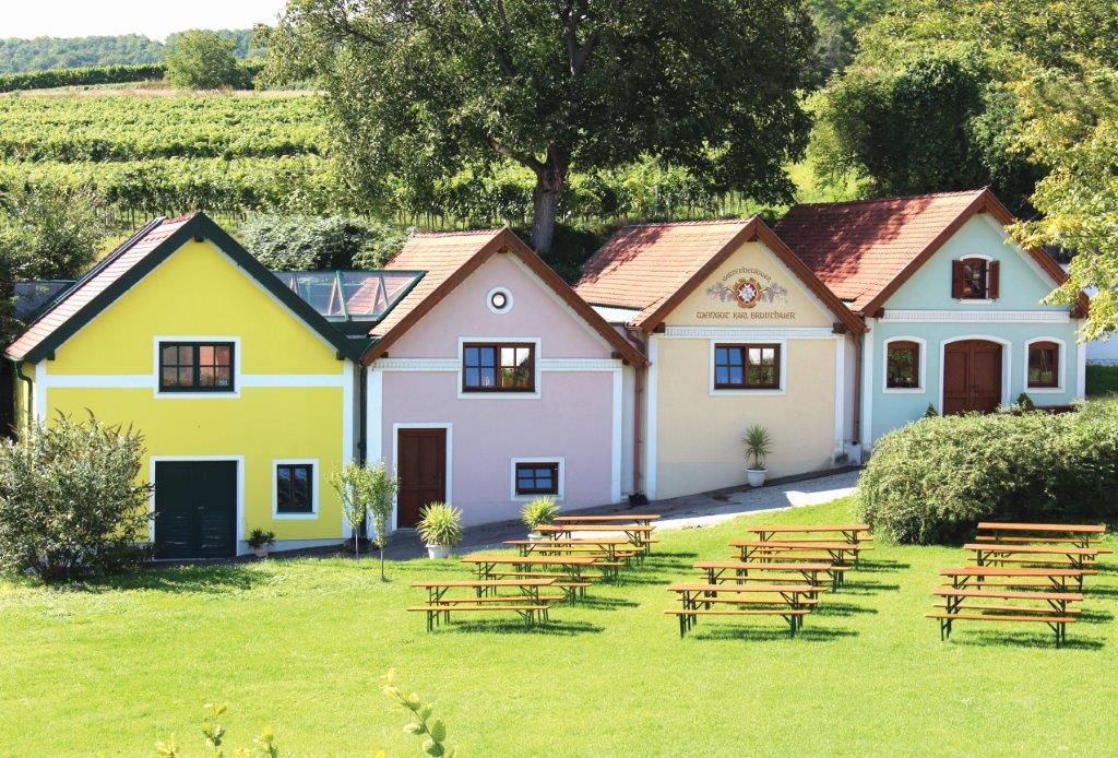 Three colorful buildings of the Brunthaler winery with beer benches in the foreground and vineyards in the background.