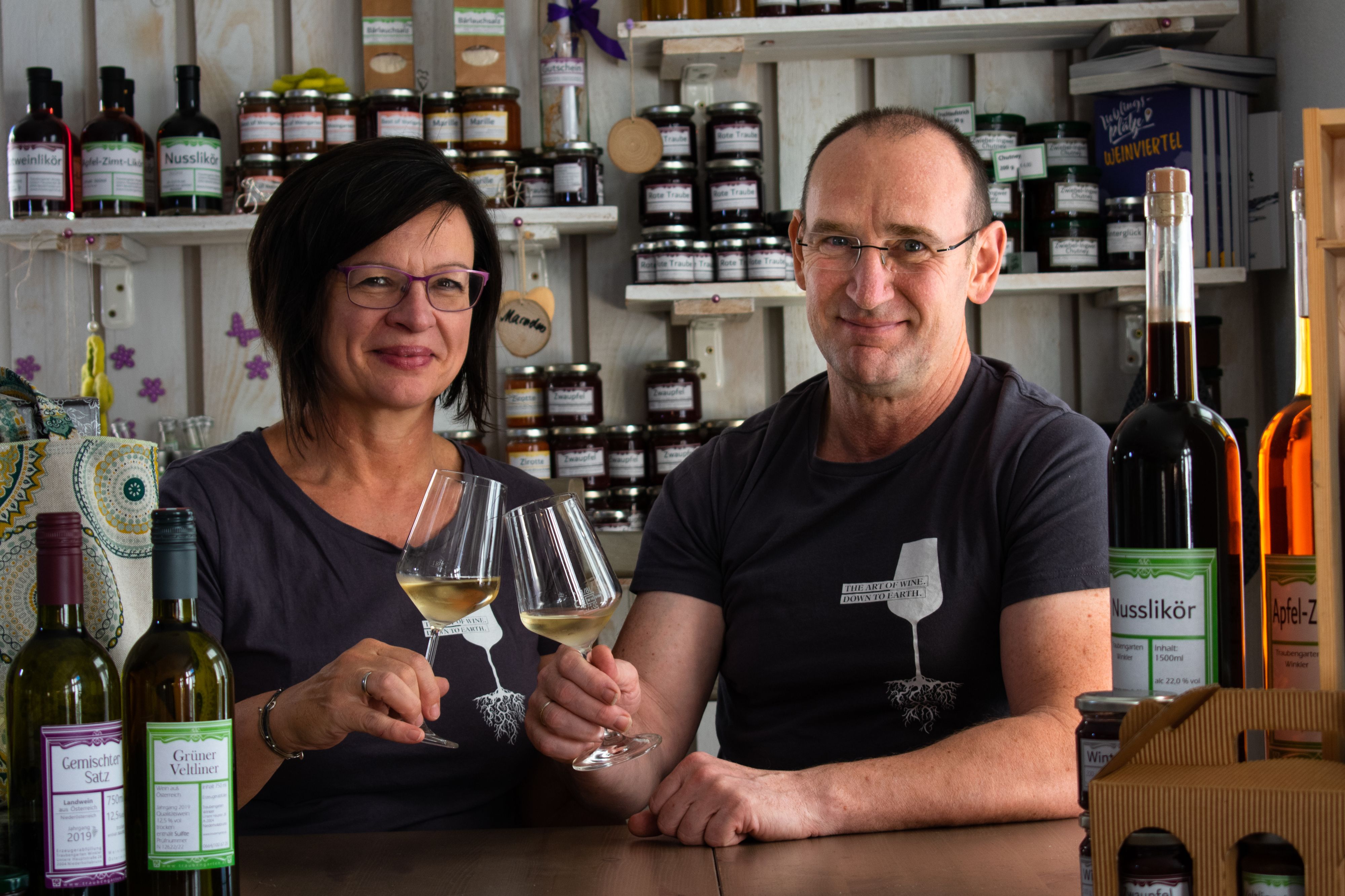 A man and a woman sit at a table in a farm store, holding glasses of wine and smiling. Shelves with products can be seen in the background.