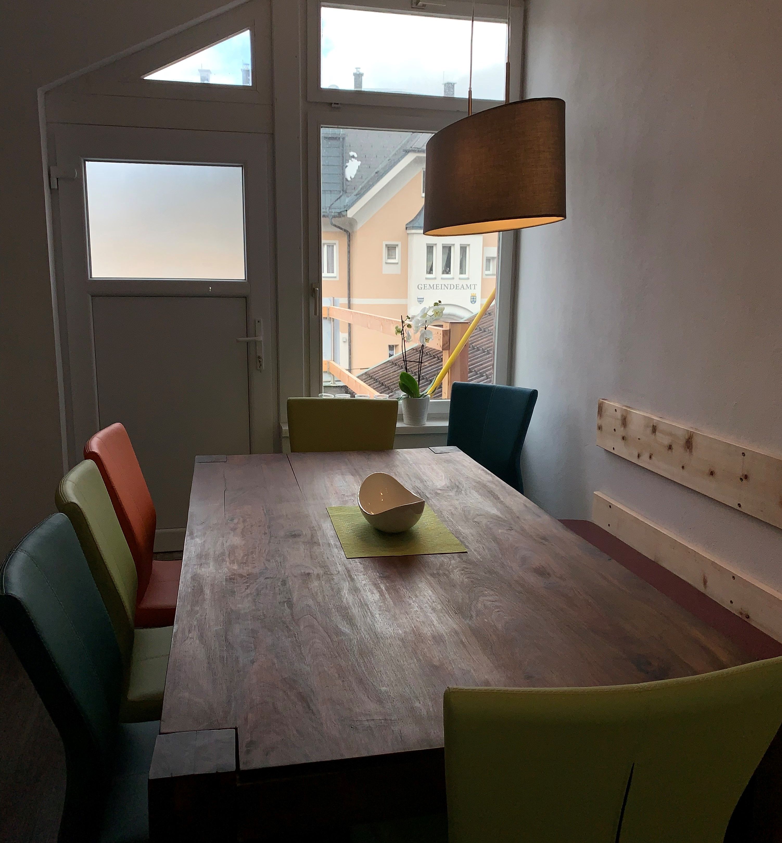 Dining area with wooden table, colorful chairs and hanging lamp, window with view of the building.