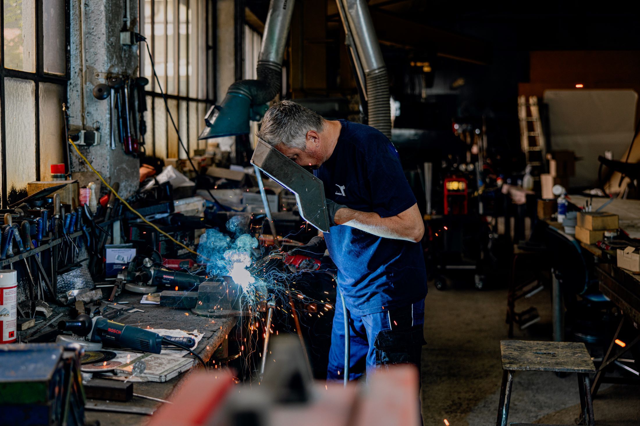 A man is welding in a workshop, surrounded by tools and sparks.