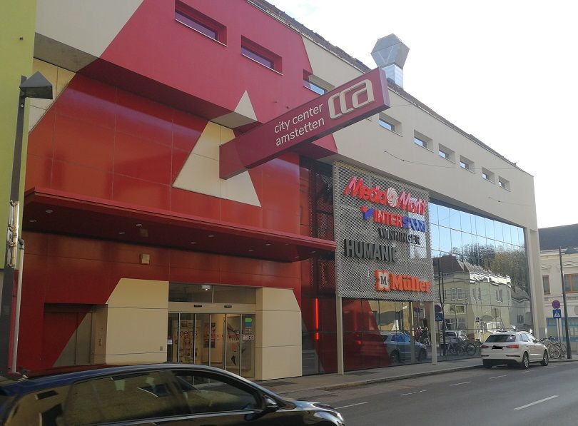 Entrance to the City Center Amstetten with red and white façade elements and signs for stores such as MediaMarkt and Müller.