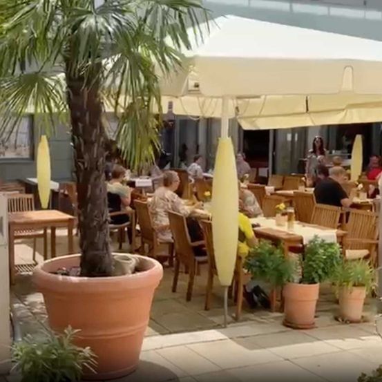 The outdoor seating area of a café with wooden tables and chairs, surrounded by plants and a large umbrella.