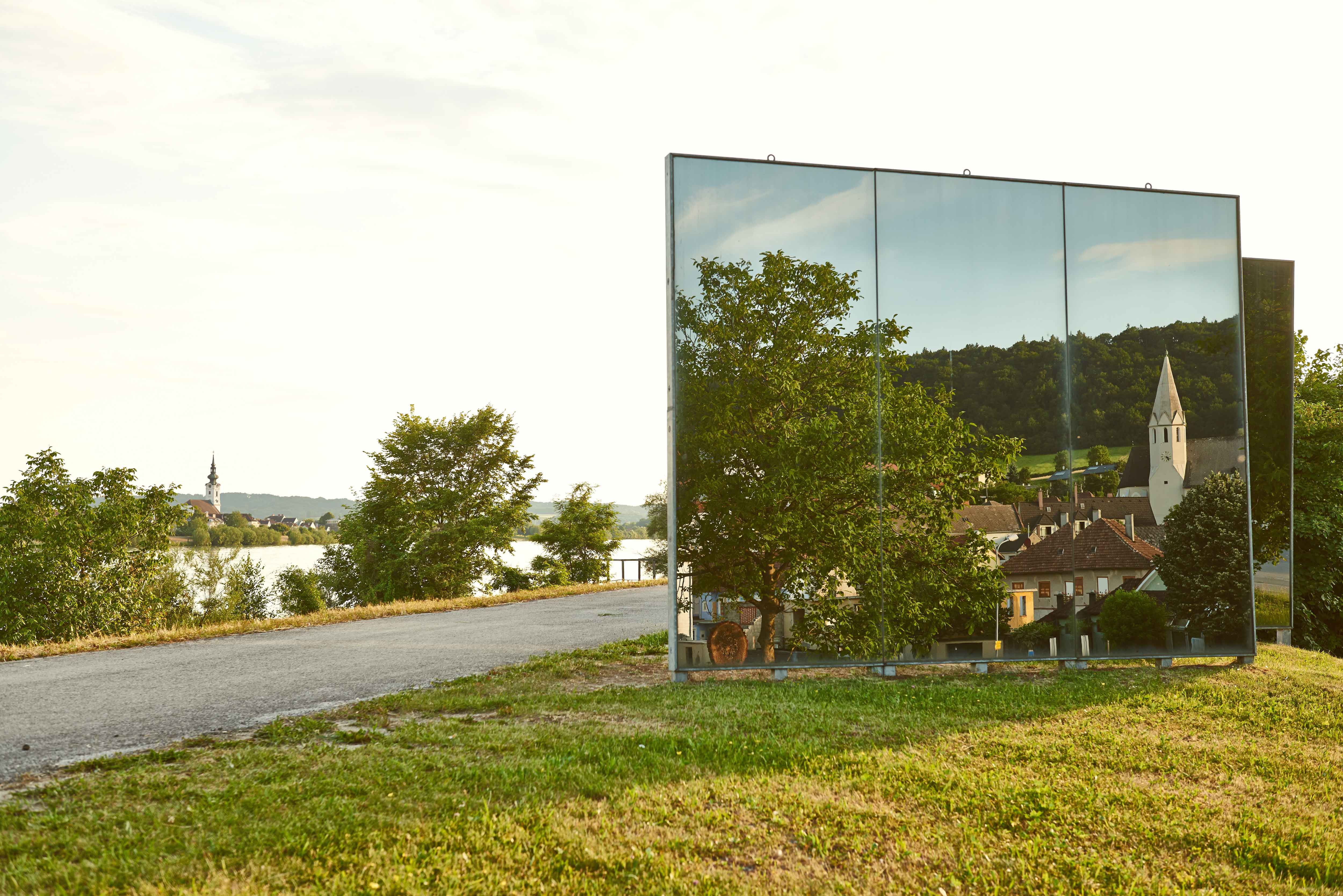 Mirror sculpture reflects landscape with church and houses.